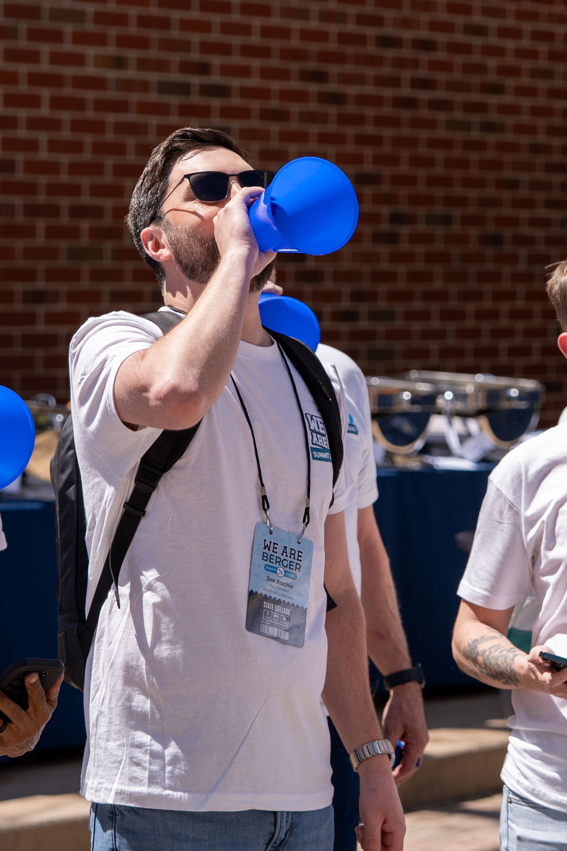 Person in sunglasses blows a blue vuvuzela at an outdoor event, wearing a white shirt and lanyard.