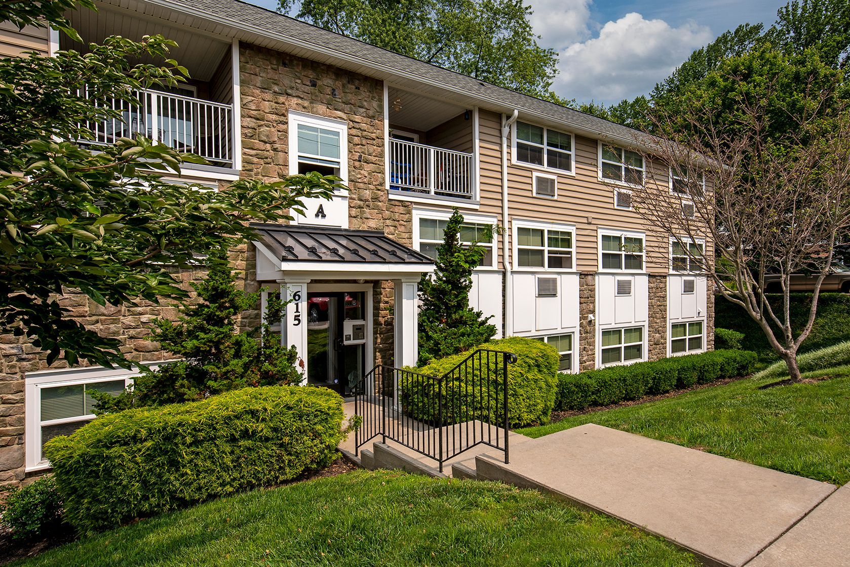 Brick apartment building with white trim, front steps, green hedges, and a sidewalk on a sunny day