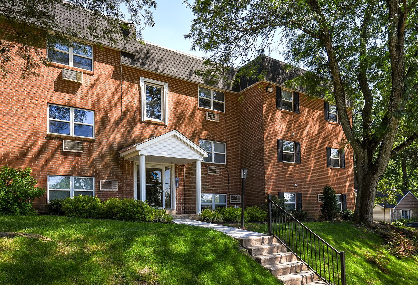 Brick apartment building with white entrance, green lawn, and stone steps under trees