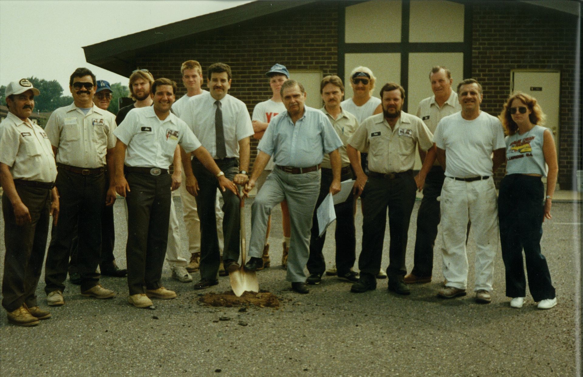 A group of people standing outside a brick building for a ground-breaking ceremony, with one person holding a shovel.