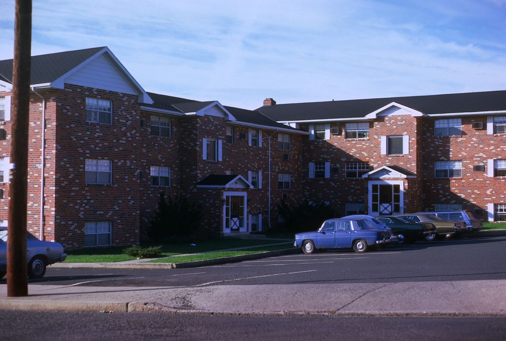 Three-story brick apartment buildings with gabled roofs, featuring several parked cars in the asphalt lot out front.