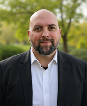Smiling bald man in a black blazer and white shirt, standing outdoors with blurred green trees in the background