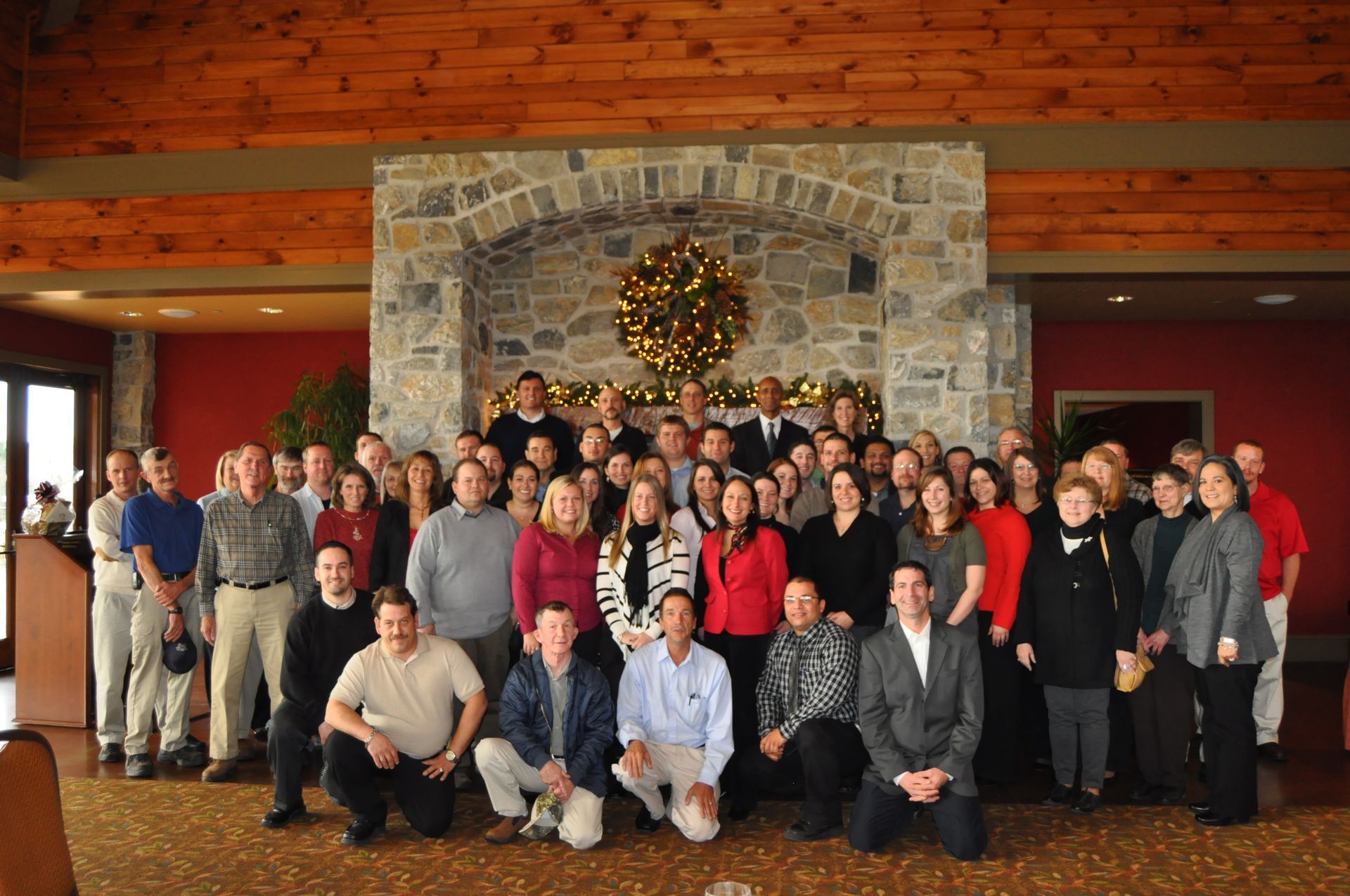 A large, diverse group of people pose for a photo in a rustic room with a stone fireplace and a holiday wreath.