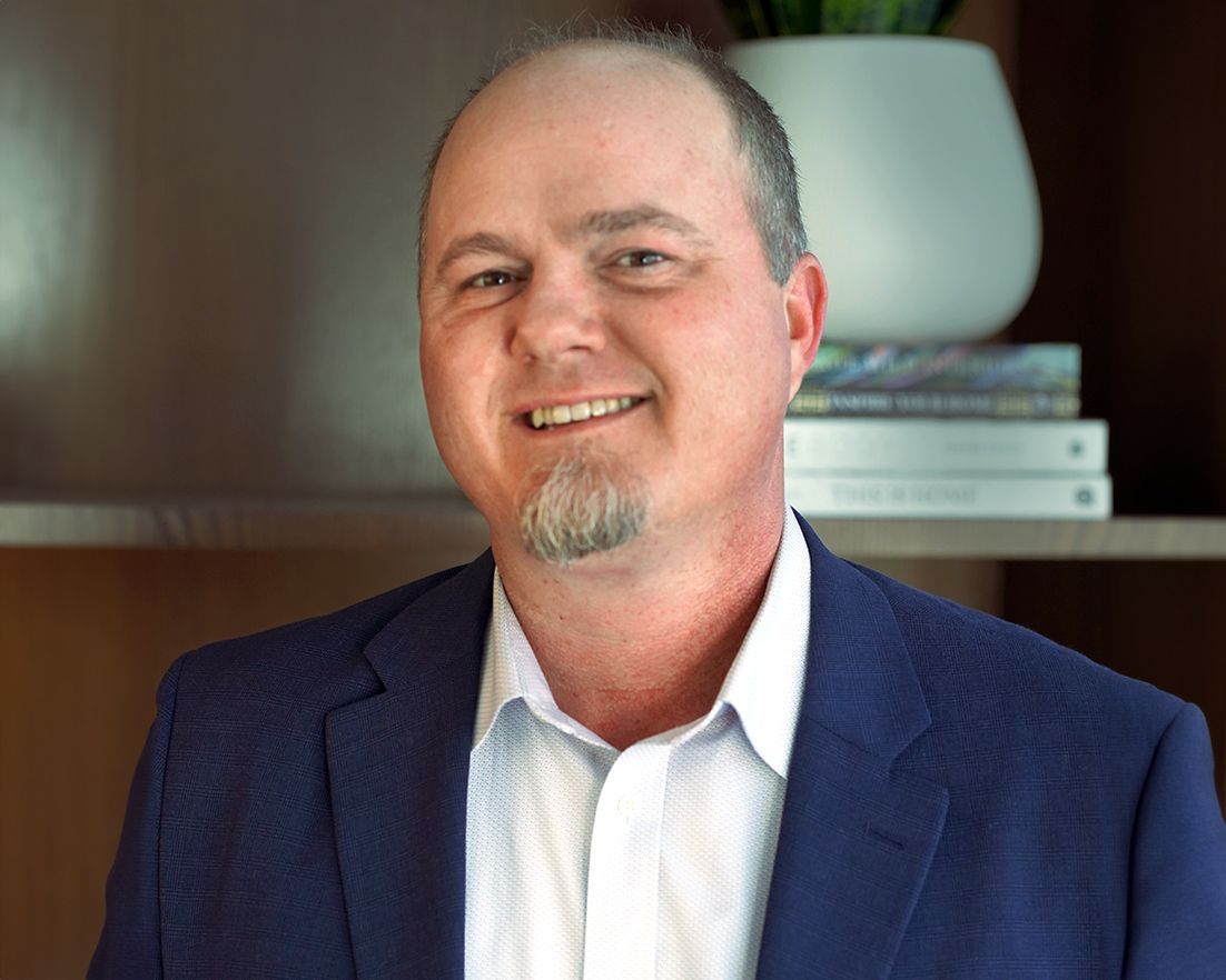 Man in blue suit, smiling with a goatee, standing in front of shelves with books and decor.