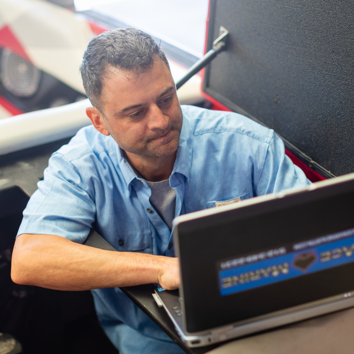 Man using a laptop, possibly for marine diagnostics. He's in a vehicle, wearing a blue shirt.