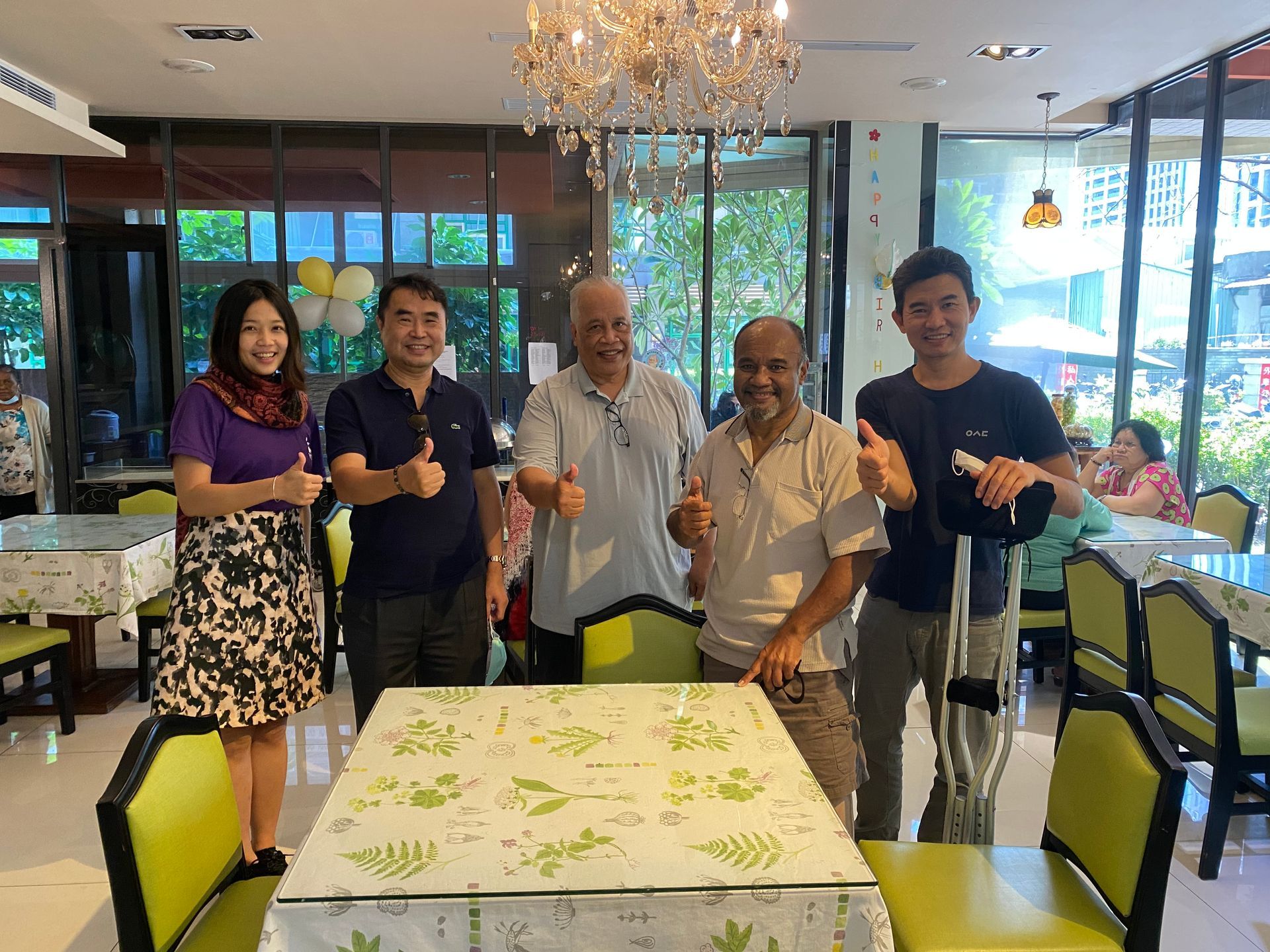 照片中，五人（兩位女性，三位男性）並排站立在一間明亮餐廳的餐桌後方，皆微笑並對著鏡頭豎起大拇指。最左側的女性穿著紫色襯衫和花紋裙子。最右側的男性穿著黑色襯衫，手中握著腋下拐杖。中間的三位男性穿著便服。