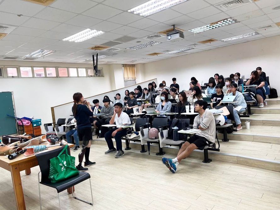 大眾教育講座現場:一名穿戴義肢的專業講師在階梯教室與學生分享,桌面上擺放多款義肢零組件,推動多元包容的友善環境。