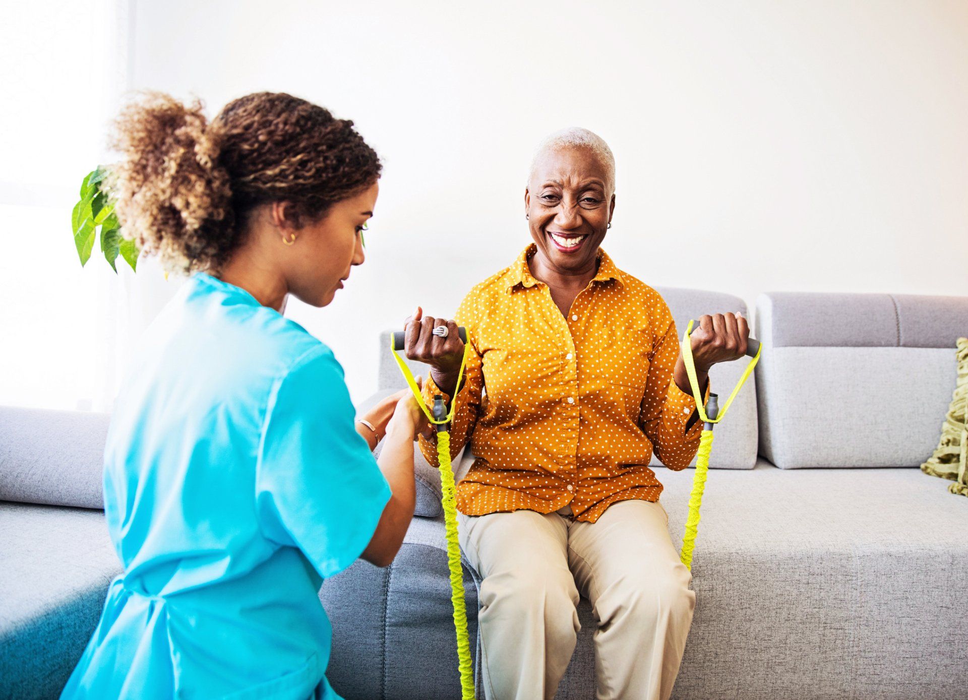 Woman using resistance bands, guided by a nurse on a couch. Both smiling.