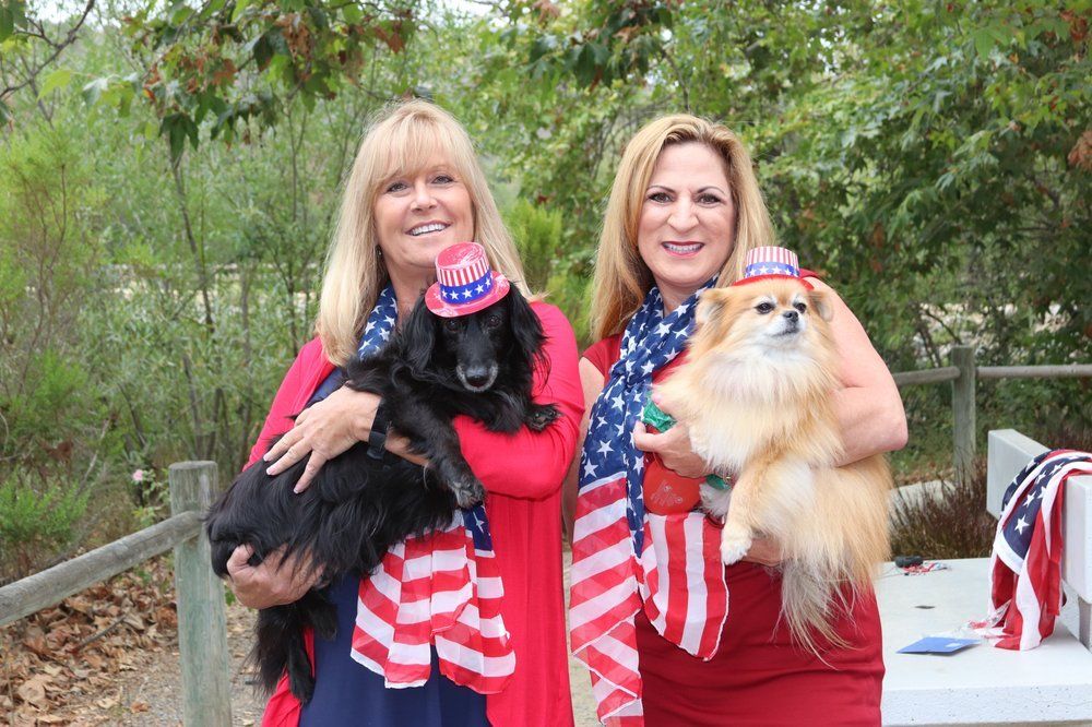 Two women, each holding a dog. They wear patriotic hats and scarves. Outdoors with foliage.