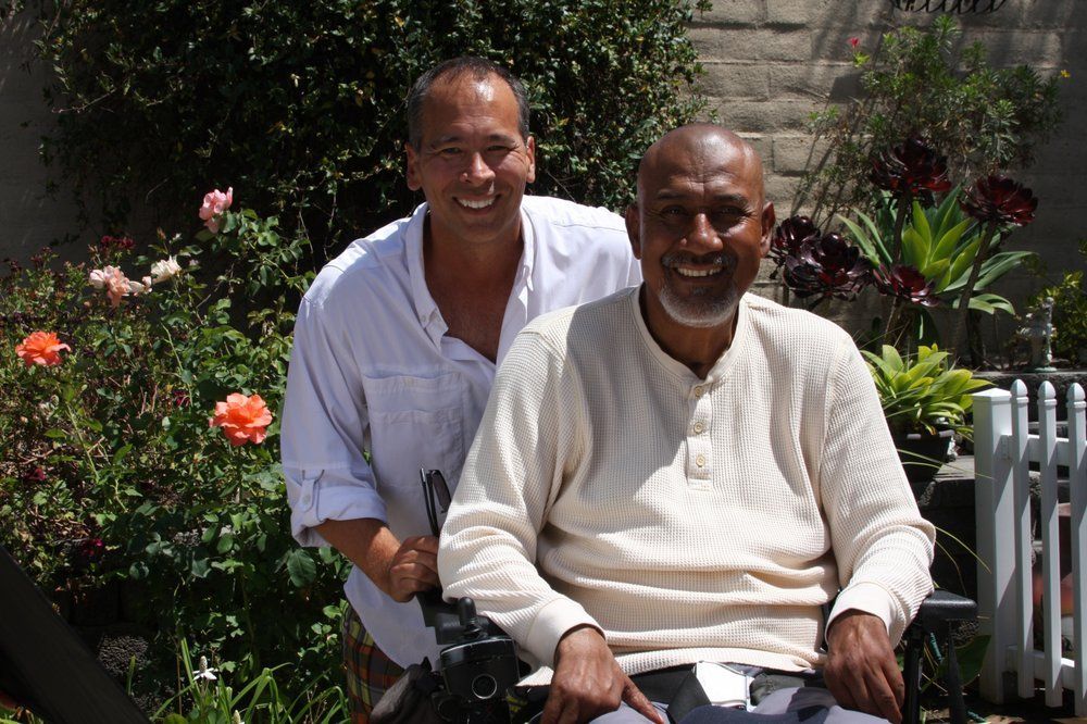 Man in wheelchair smiles at the camera, another man smiles behind him in a garden.