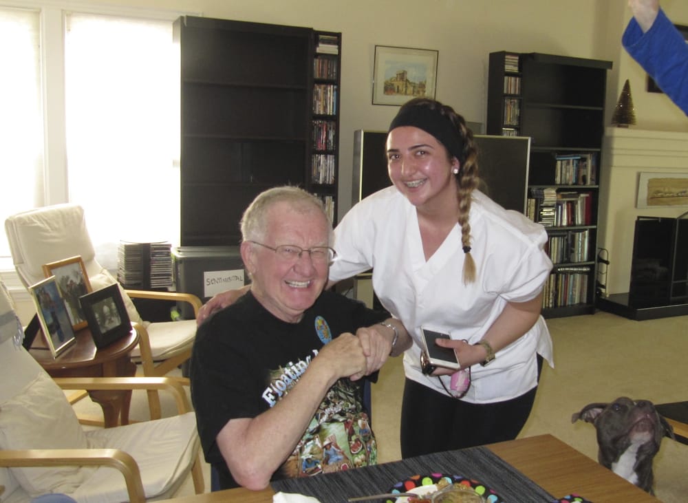 Elderly man and young woman smiling, holding hands, in a room. Man wears a graphic tee, woman wears scrubs.