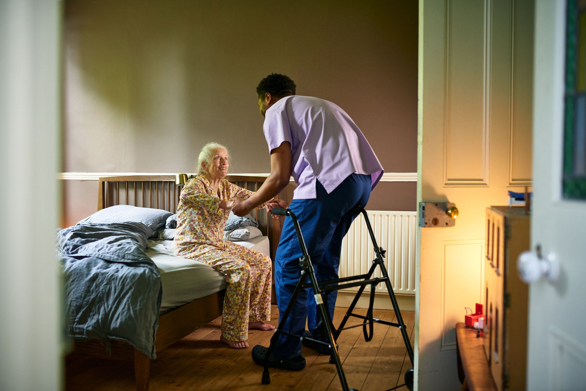 Caregiver providing mobility help as elderly woman gets up from bed.
