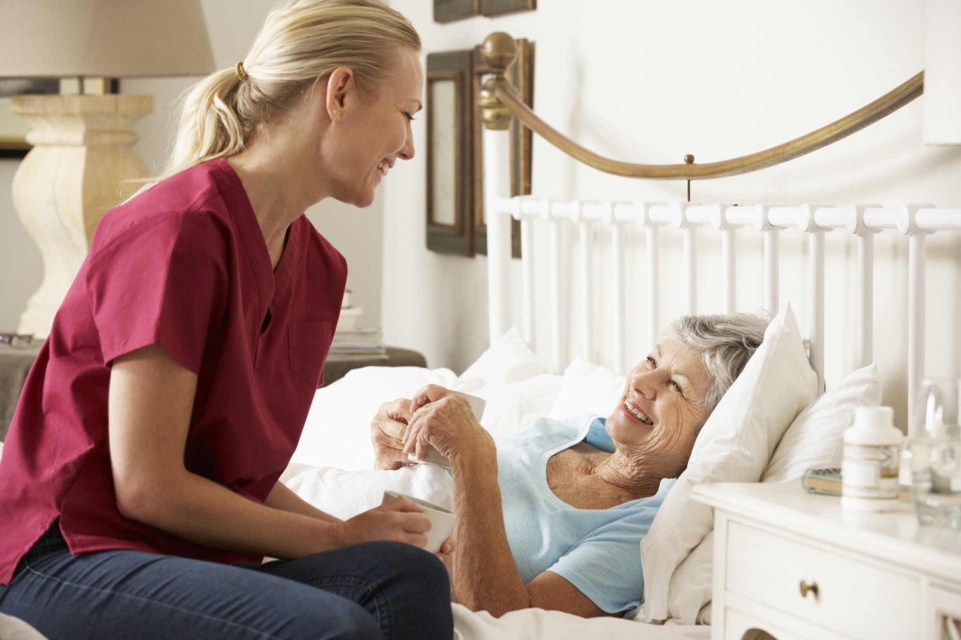 Caregiver and senior woman in bed, smiling, holding hands. Bedroom setting.