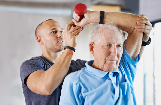 Man helps senior citizen lift a small dumbbell. They are indoors, with the older man wearing a blue shirt.