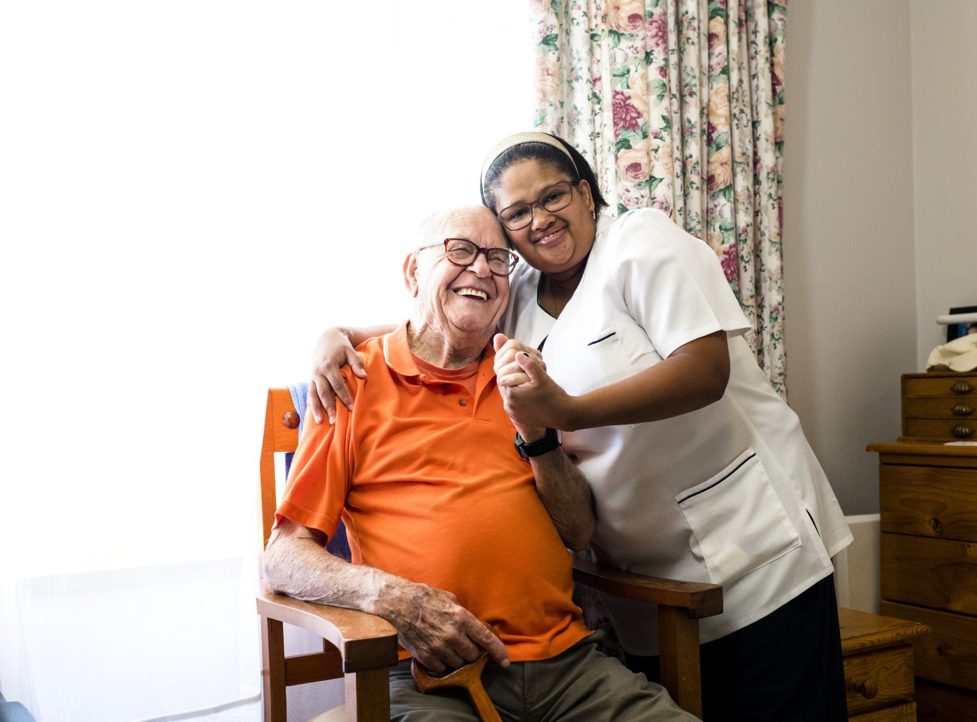 Caregiver embracing and smiling with an elderly man in a room.