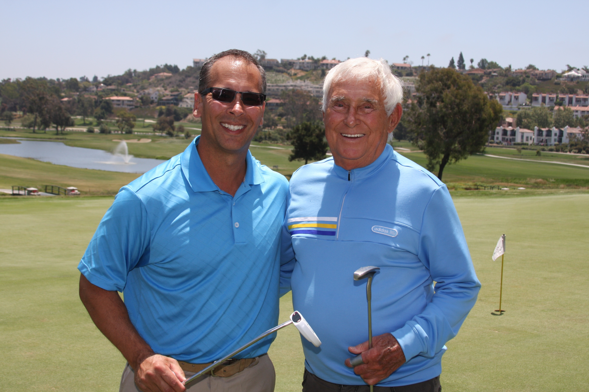 Two men in blue shirts smile on a golf course. Man on right holds a putter. Green landscape.