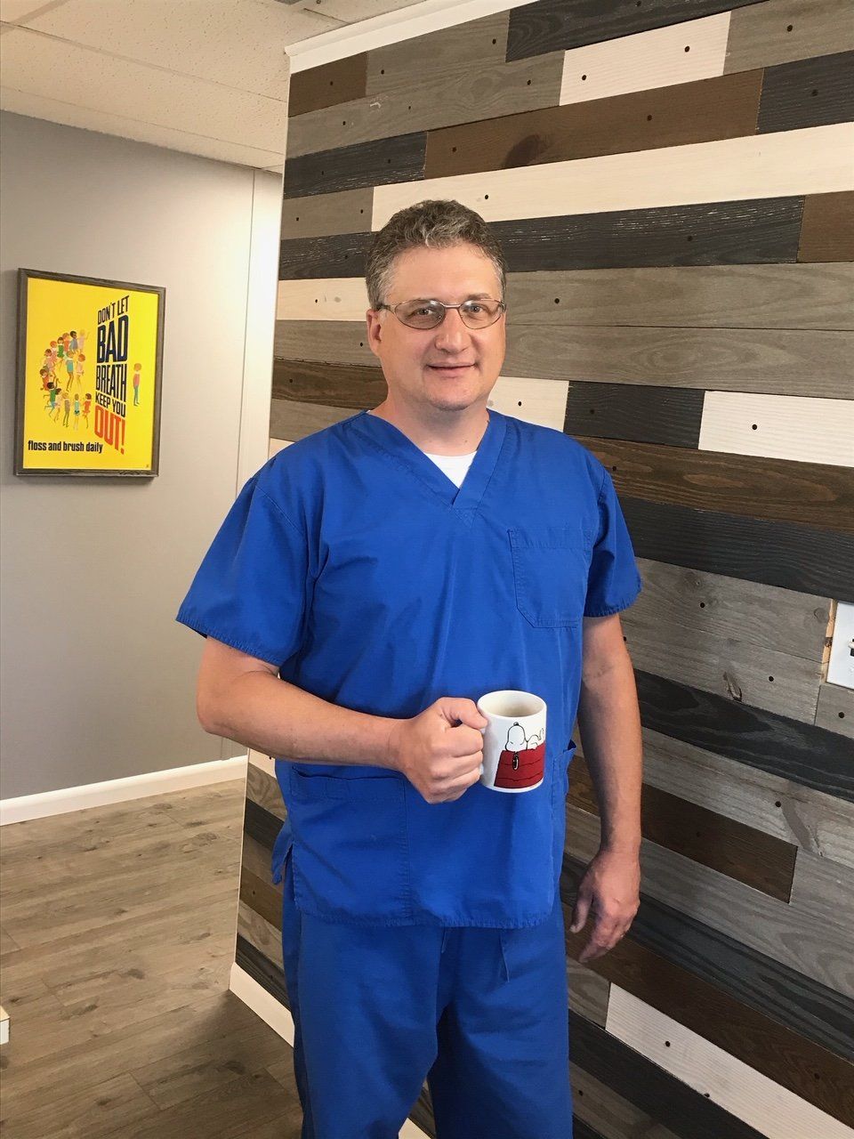 Man in blue scrubs holding a mug, standing near a wood-paneled wall, smiling.
