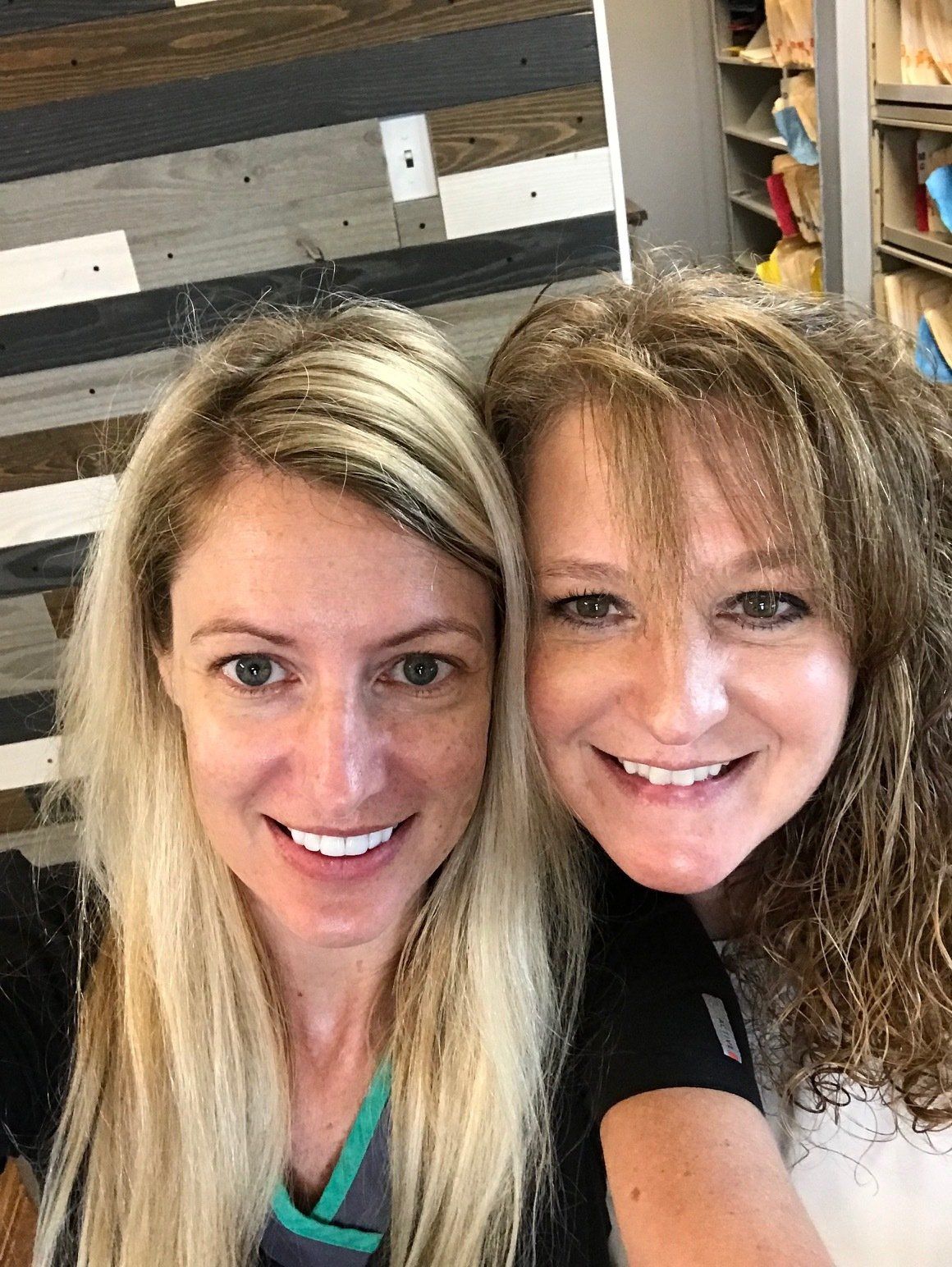 Two smiling women, close up selfie. Blonde and brunette hair, in an office setting.