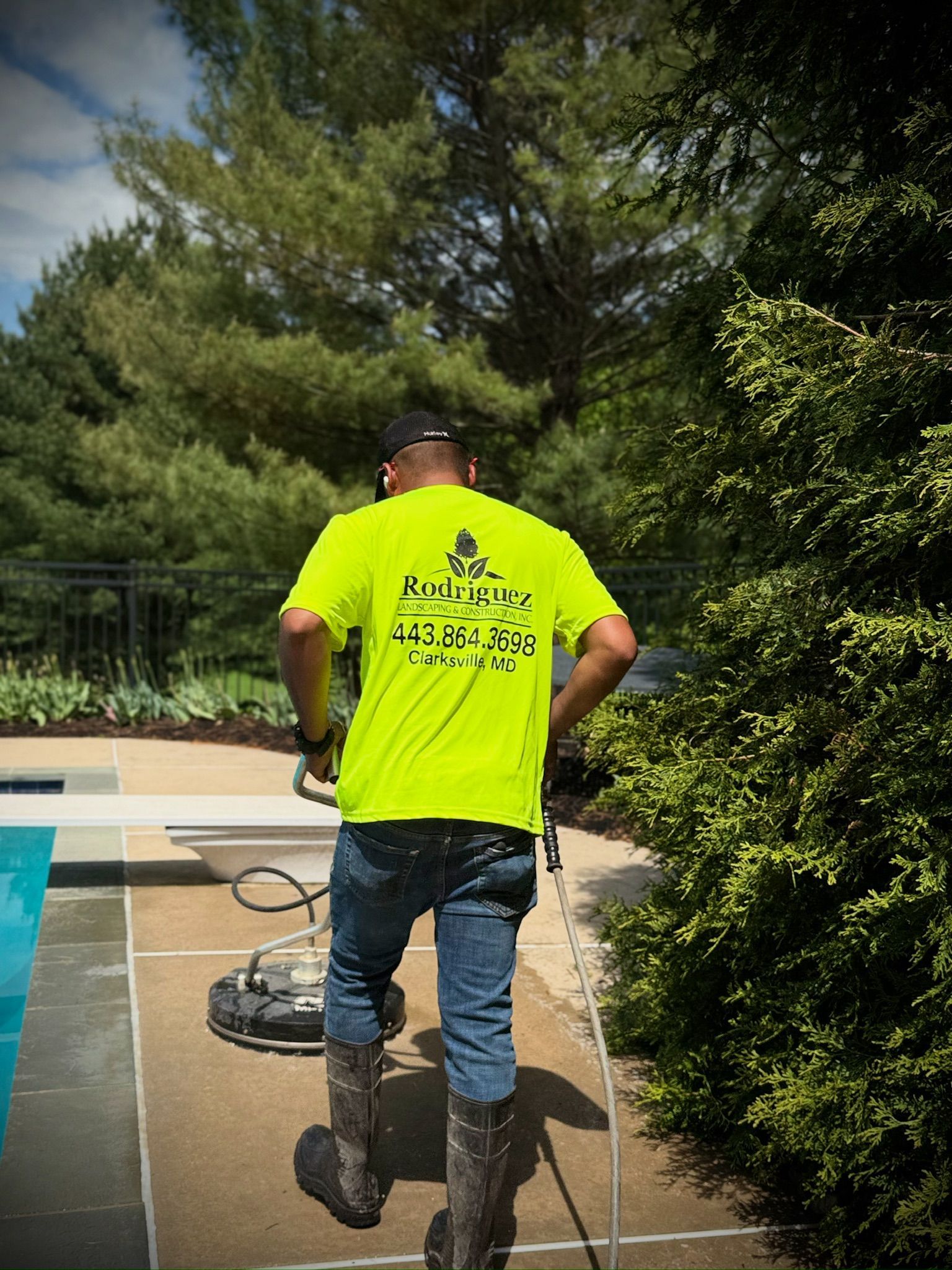 Worker in neon yellow shirt pressure-washing a pool deck beside a swimming pool.