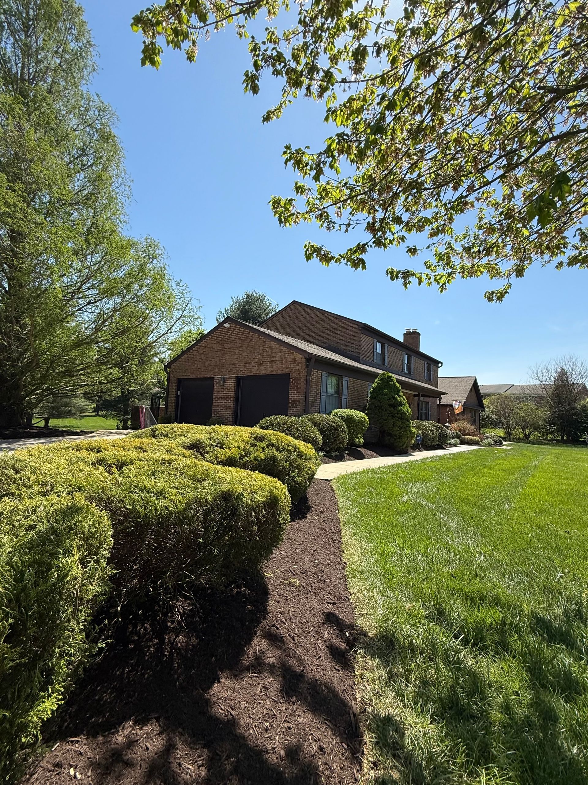 Brick house with a tiled roof beside a green lawn, hedges, and trees under a clear blue sky