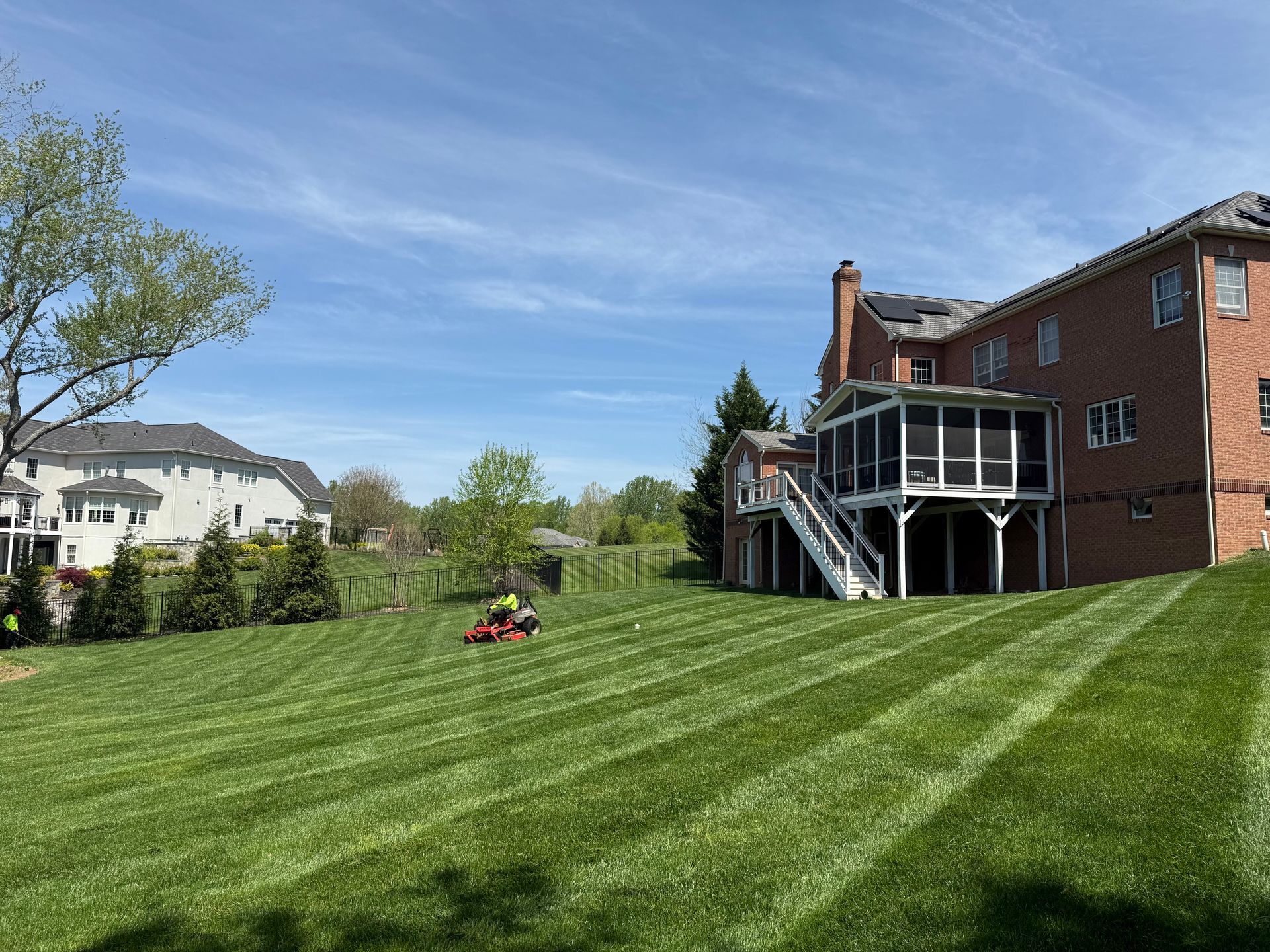 Backyard with striped green lawn, lawn mower, and brick house with deck under a blue sky