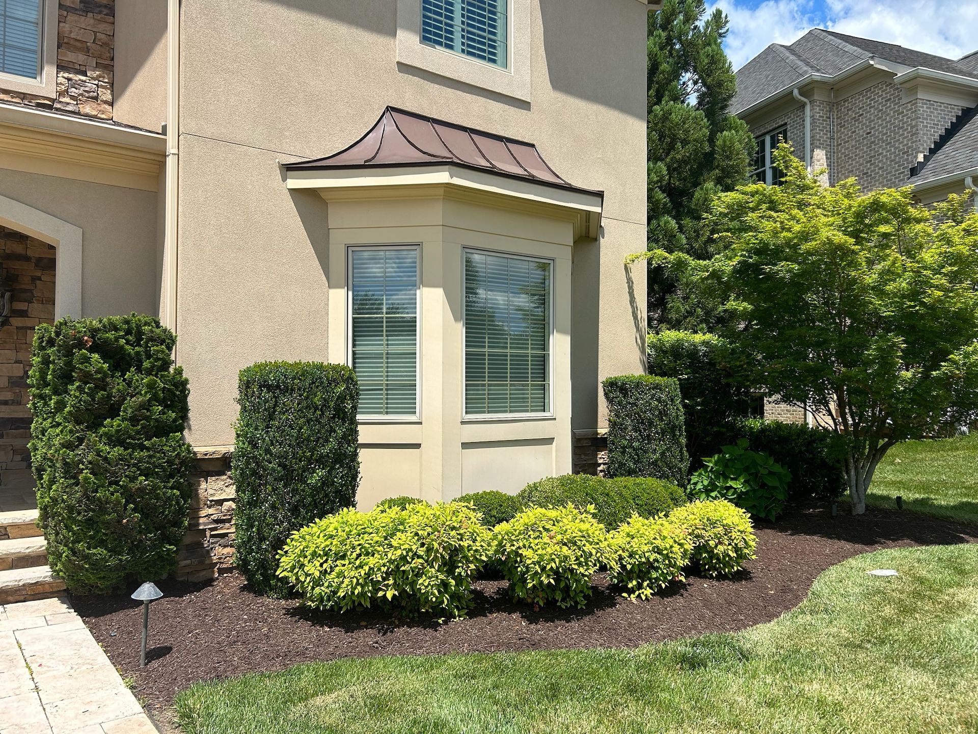 Front of a beige house with trimmed hedges, a bay window, and a landscaped garden bed.