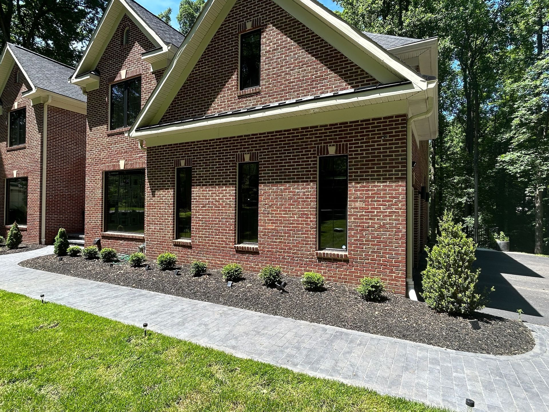 Brick house with steep gabled roof, black windows, and a gravel path bordered by shrubs.
