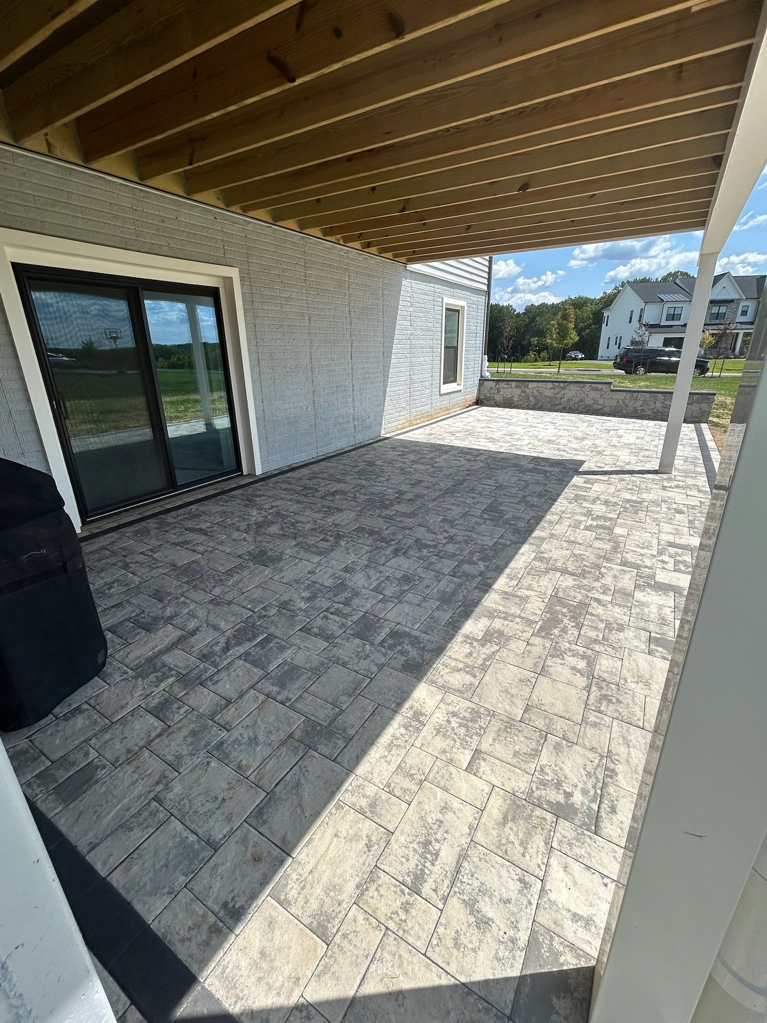 Covered patio with wood beam ceiling, stone pavers, and sliding glass doors beside a white house.