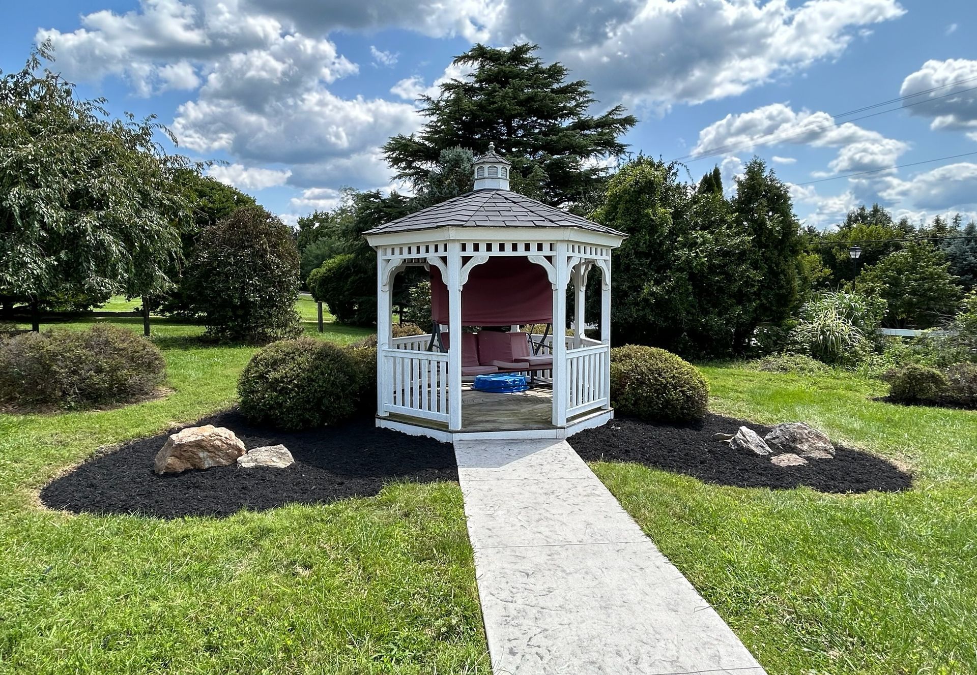 White gazebo on a lawn with a stone path, surrounded by shrubs under a sunny blue sky.