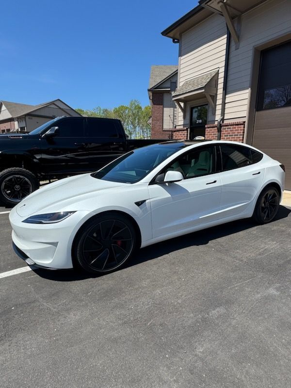 White Tesla sedan parked in a driveway beside a black pickup truck and a house