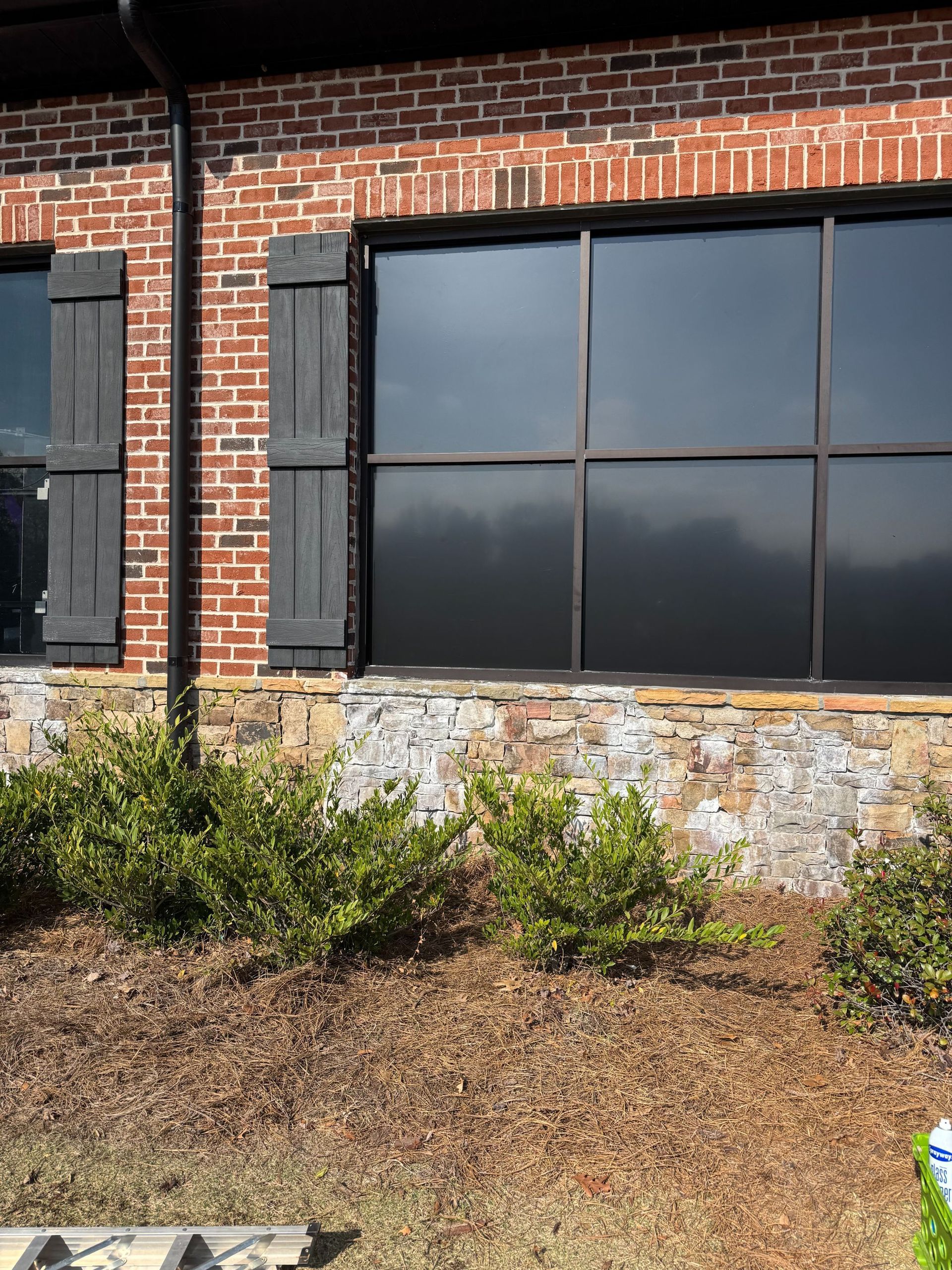 Brick building exterior with large dark windows, stone base, and shrubs in front
