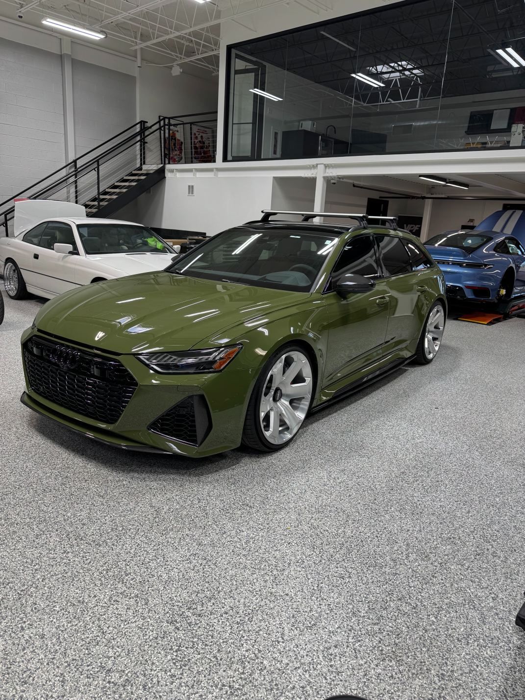 Olive green sports sedan in a showroom, with other cars parked behind it