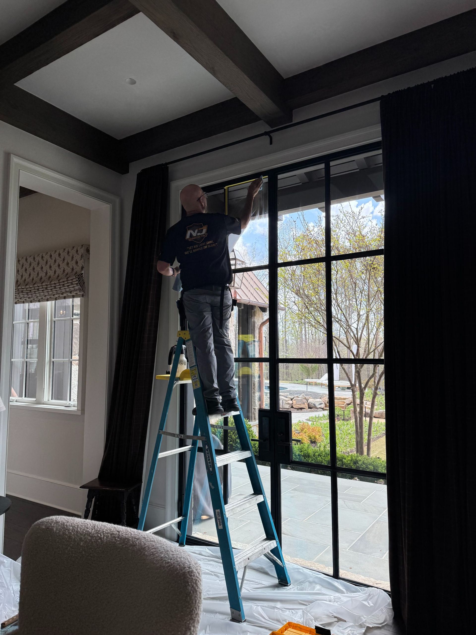 Person on a ladder hanging black-framed glass doors in a modern room with exposed ceiling beams.