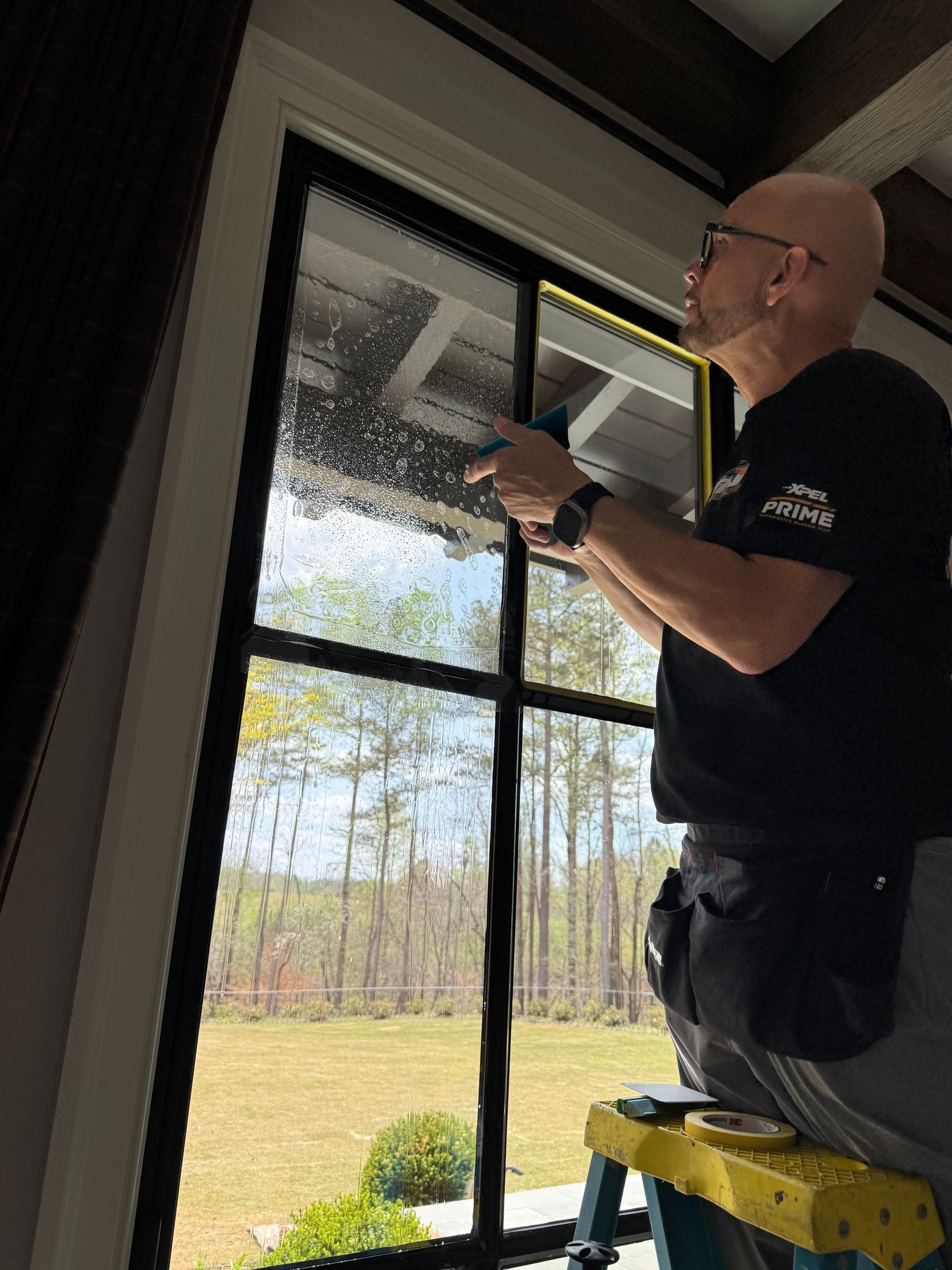 Worker on a ladder adjusting a window screen inside a bright room.