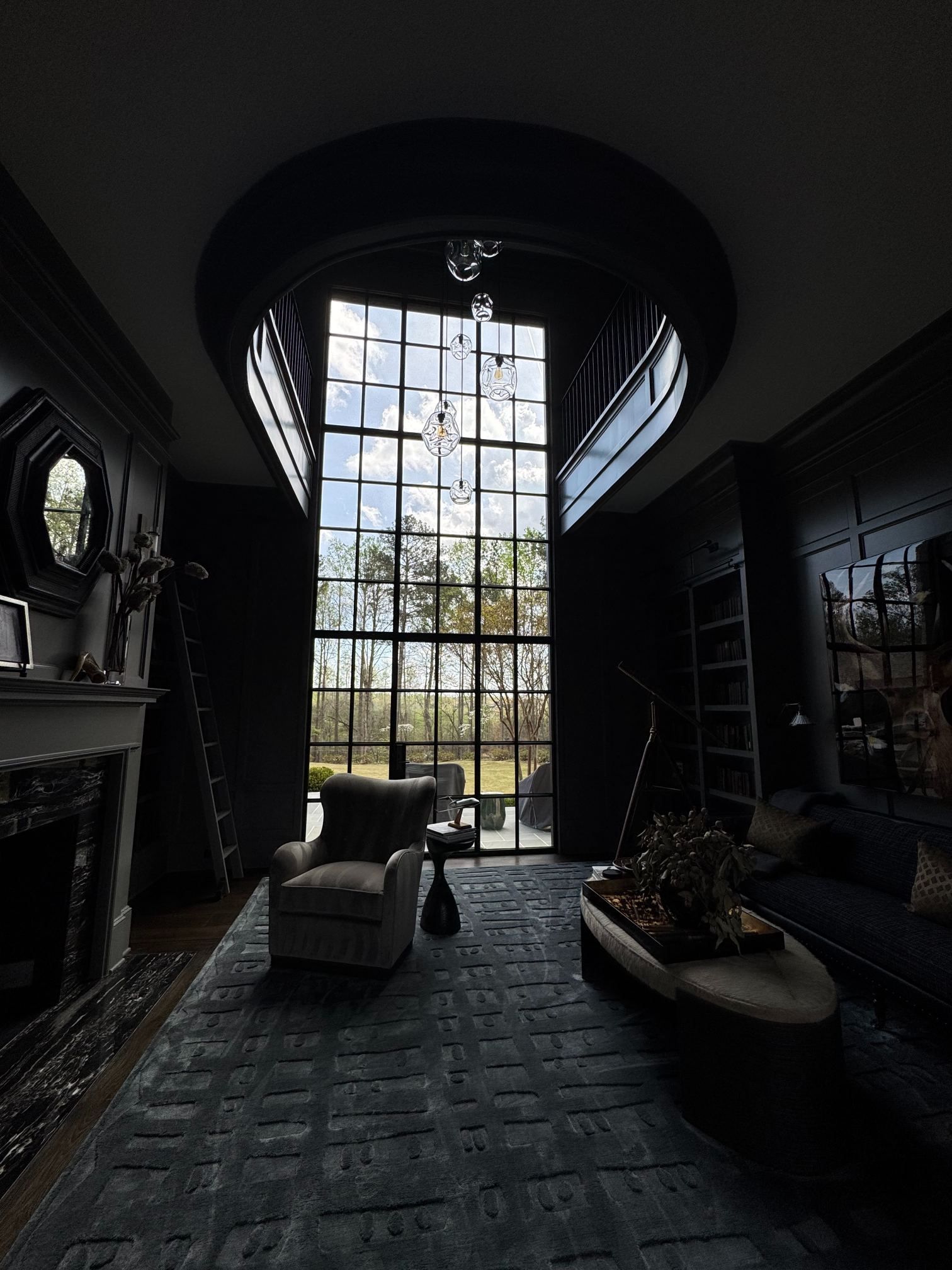 Dim modern living room with a tall arched window, two chairs, a sofa, and a hanging chandelier.