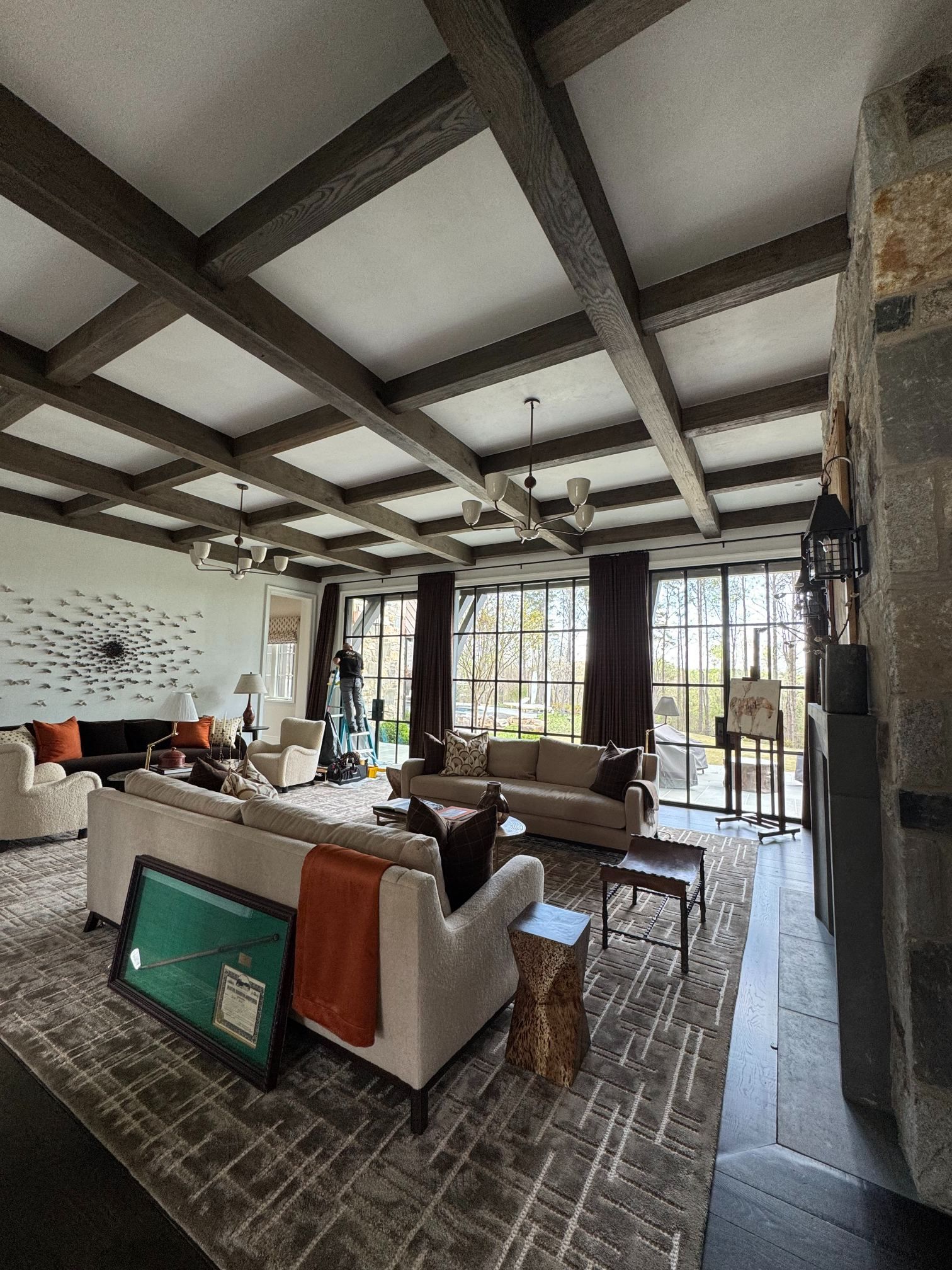 Modern living room with exposed beams, sofas, rug, fireplace, and large windows overlooking greenery