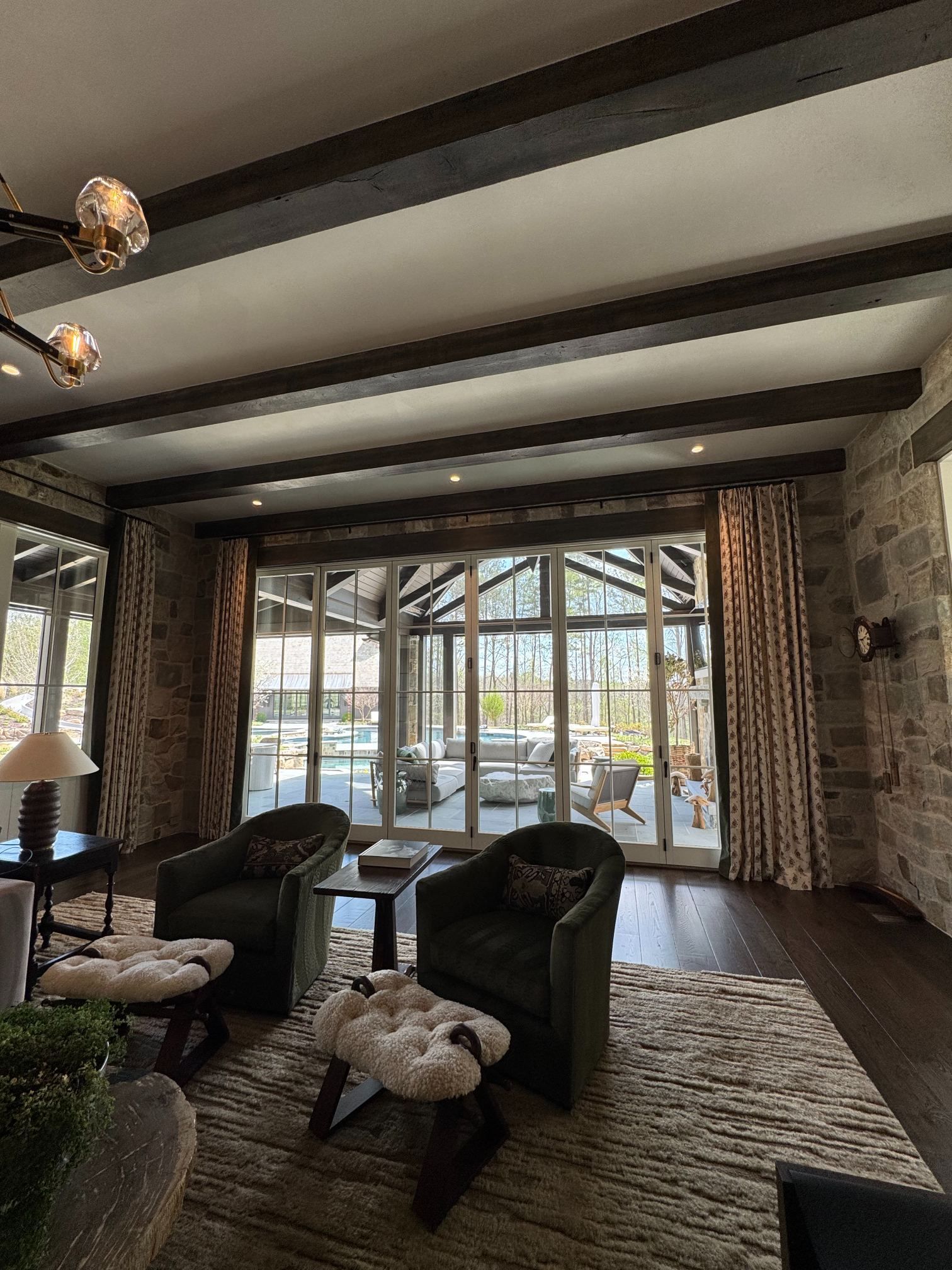 Living room with dark wood beams, green chairs, rug, and large windows overlooking a patio