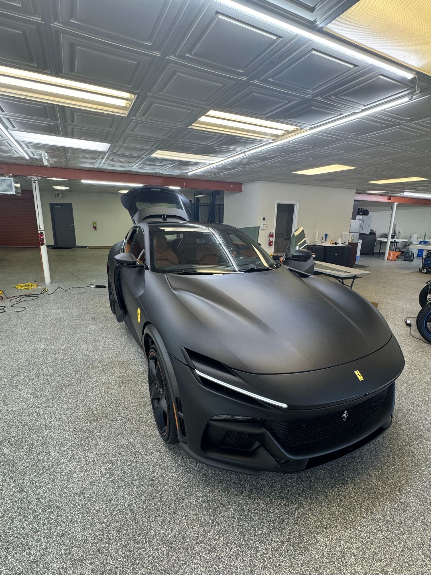 Matte black sports car in a garage, front view under bright ceiling lights.