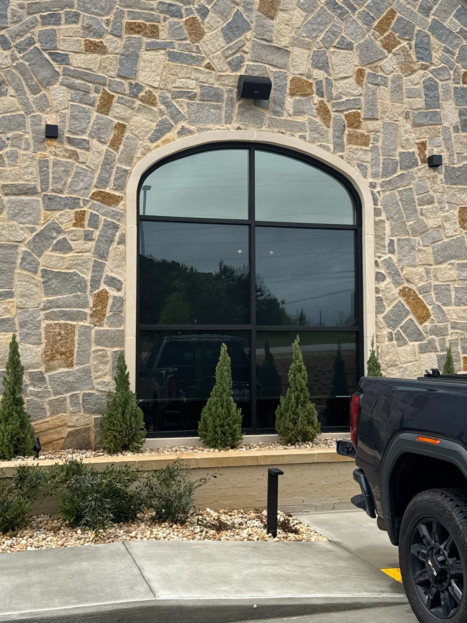 Stone building facade with arched black window, shrubs, and a parked black truck at the curb