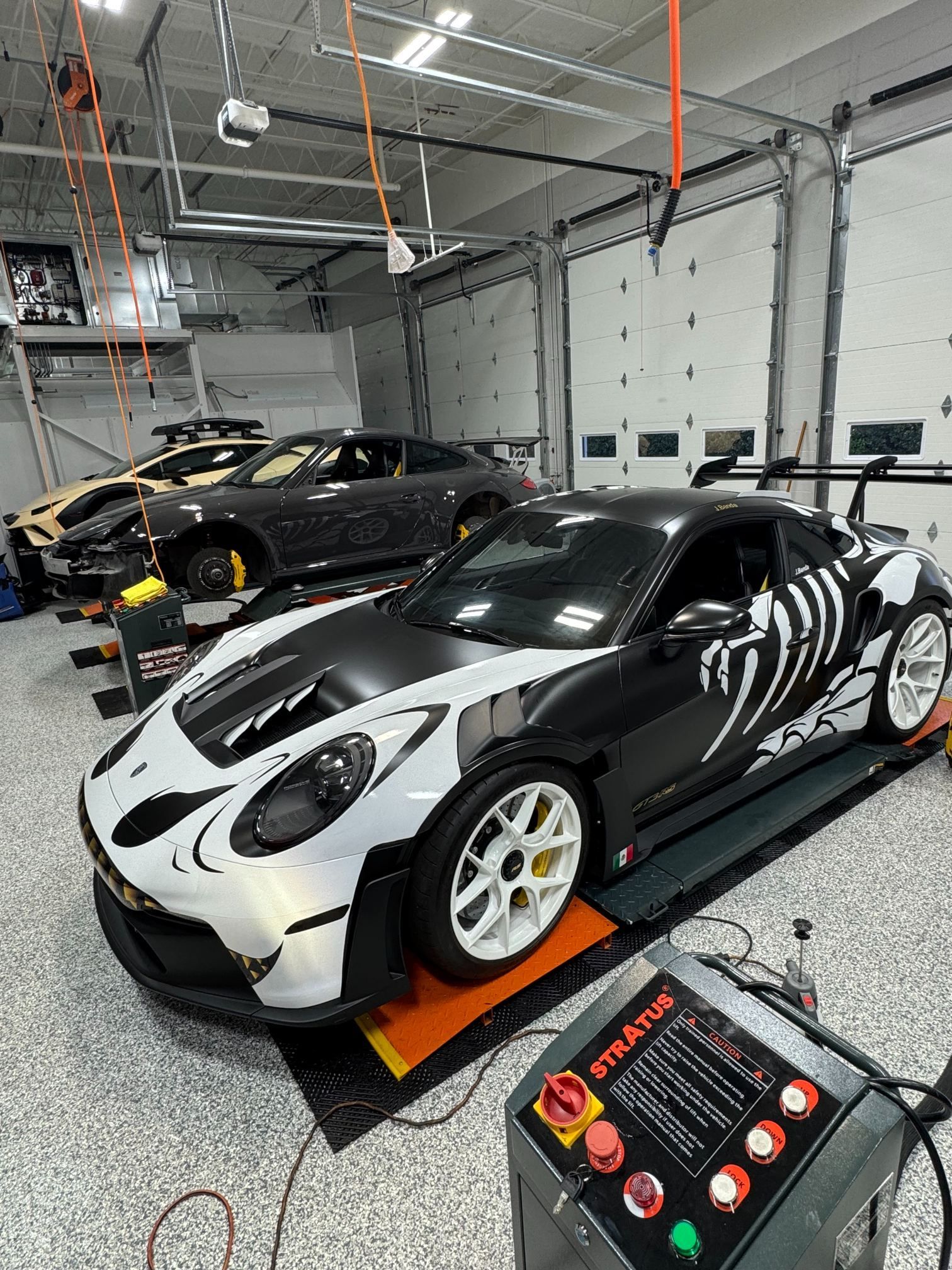 Two sports cars in a garage, with a white-and-black Porsche in the foreground on a display mat.