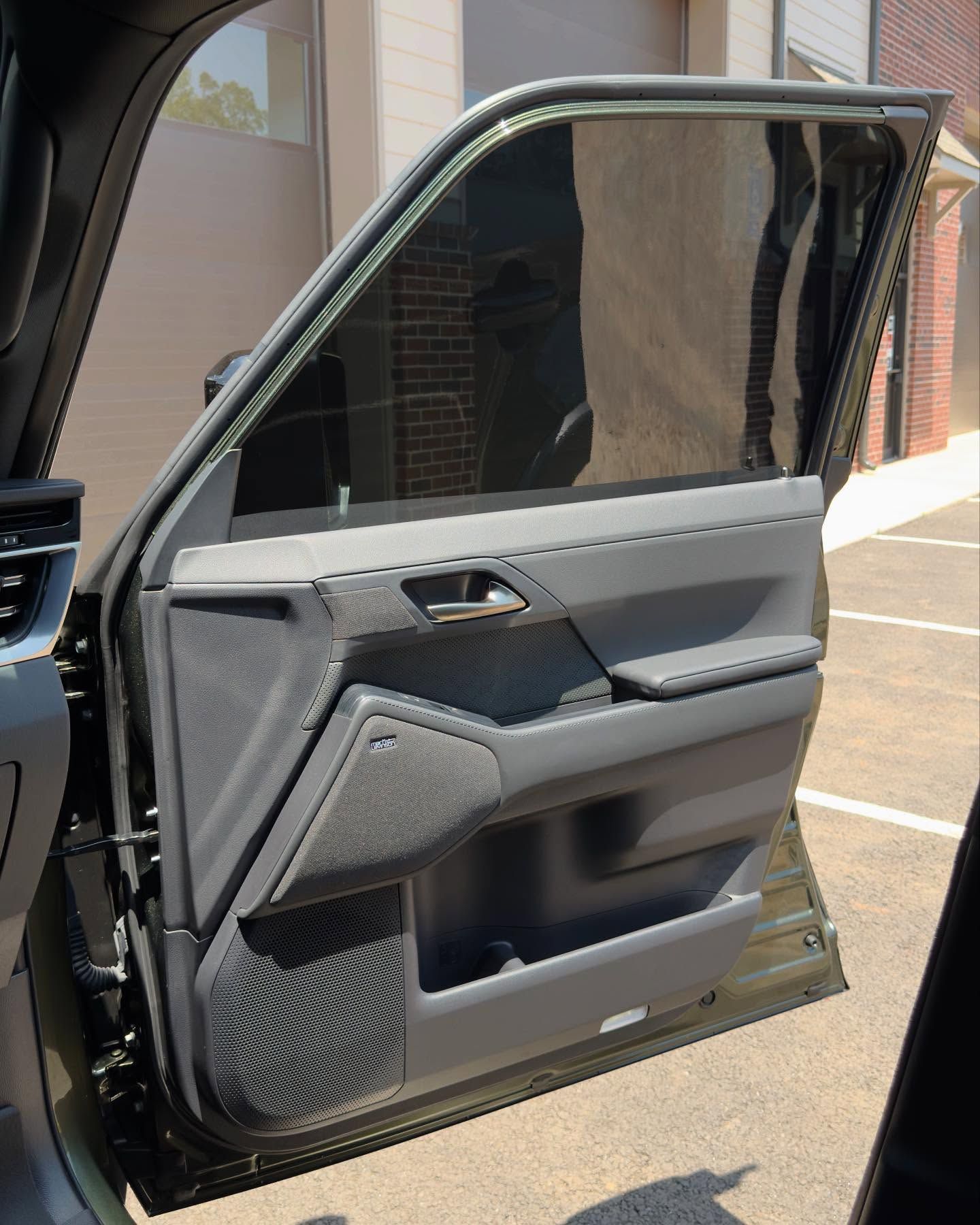 The interior panel of a dark gray car door, featuring a tinted window and a speaker, viewed from an open doorway.
