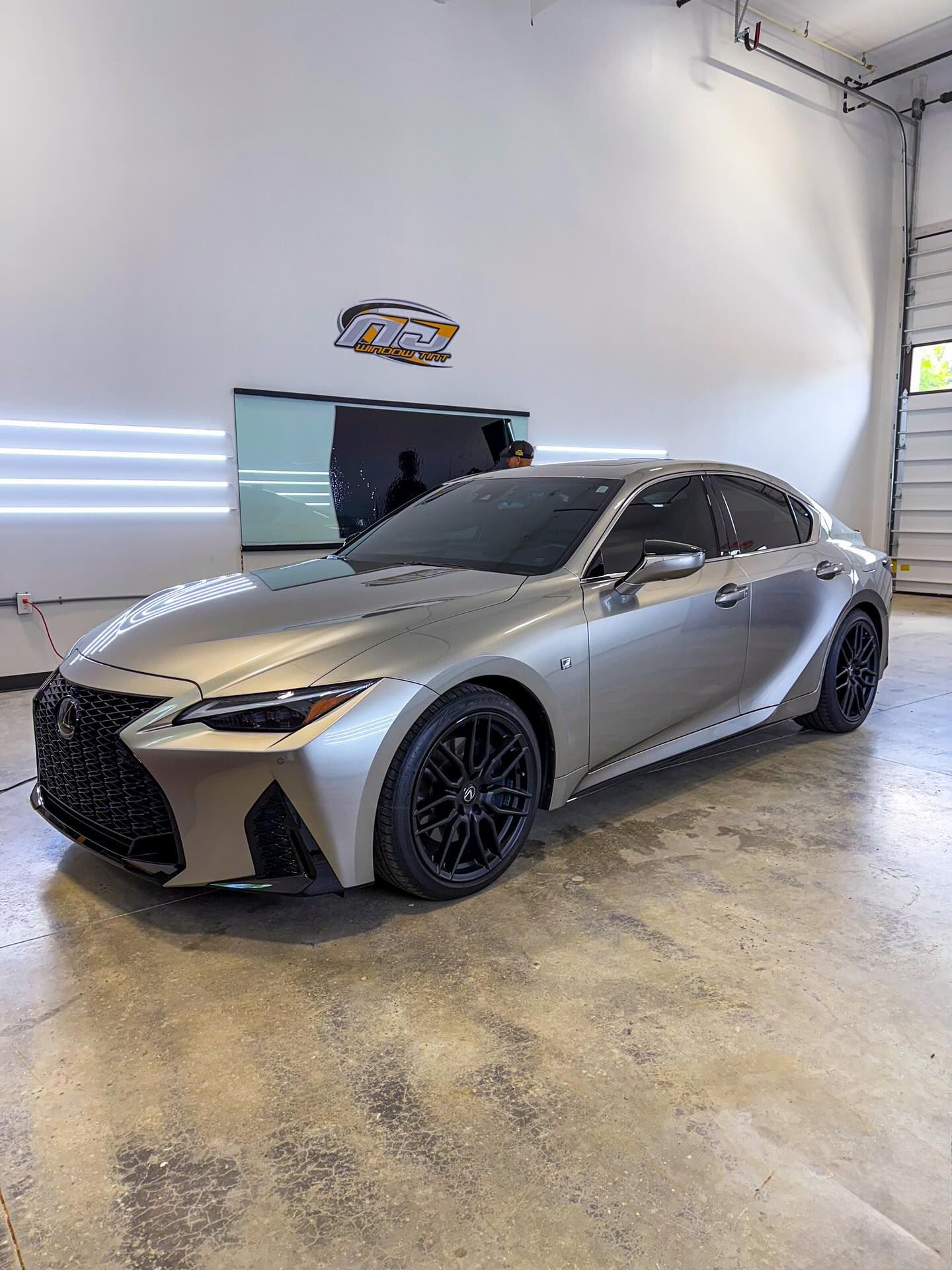 A matte silver Lexus IS sedan with black rims parked in a bright, modern auto shop with overhead lighting.