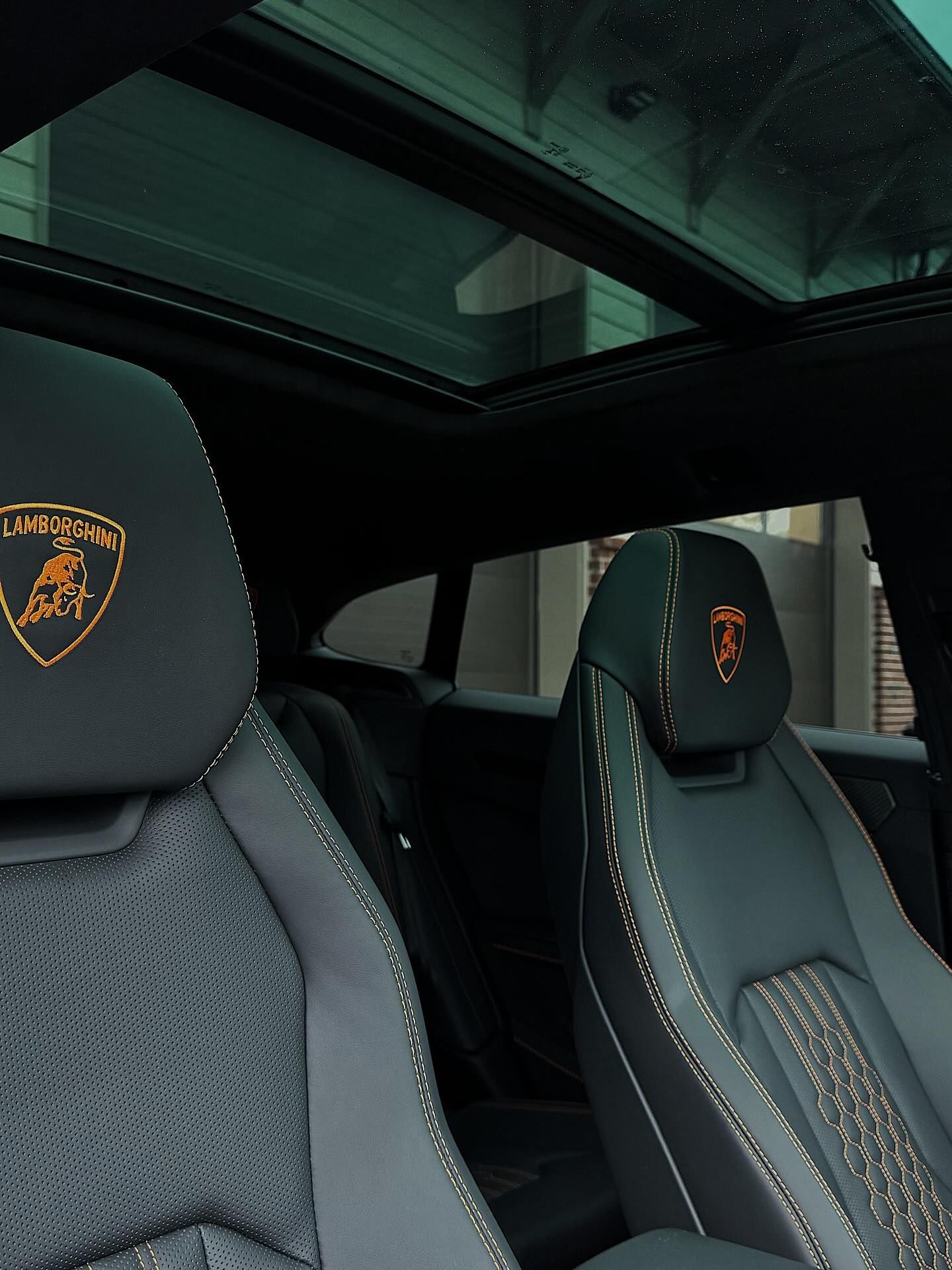 Interior of a Lamborghini with dark leather bucket seats, stitched logos, and a panoramic sunroof.