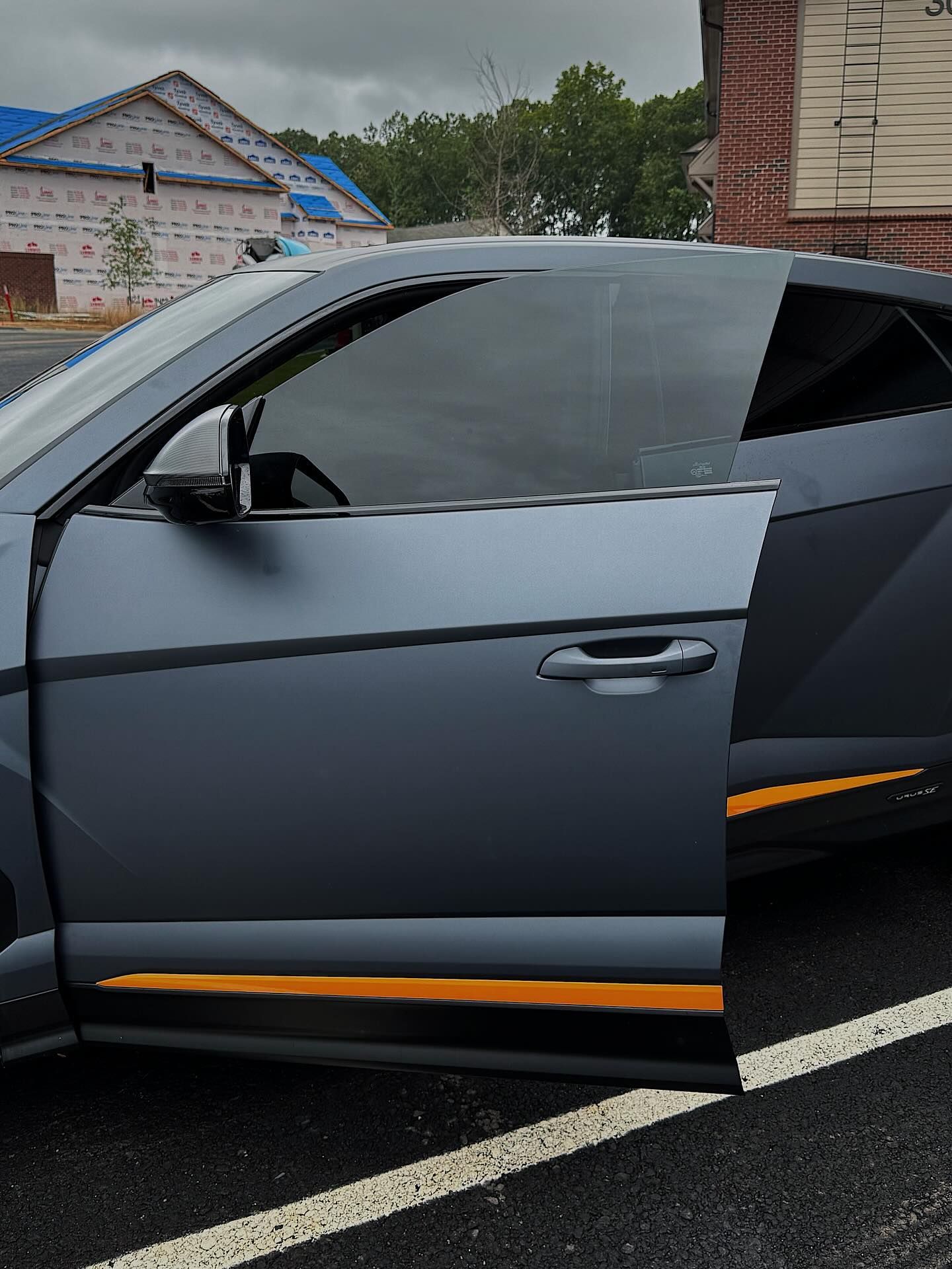 A matte grey sports car door with orange detailing stands open in a parking lot.