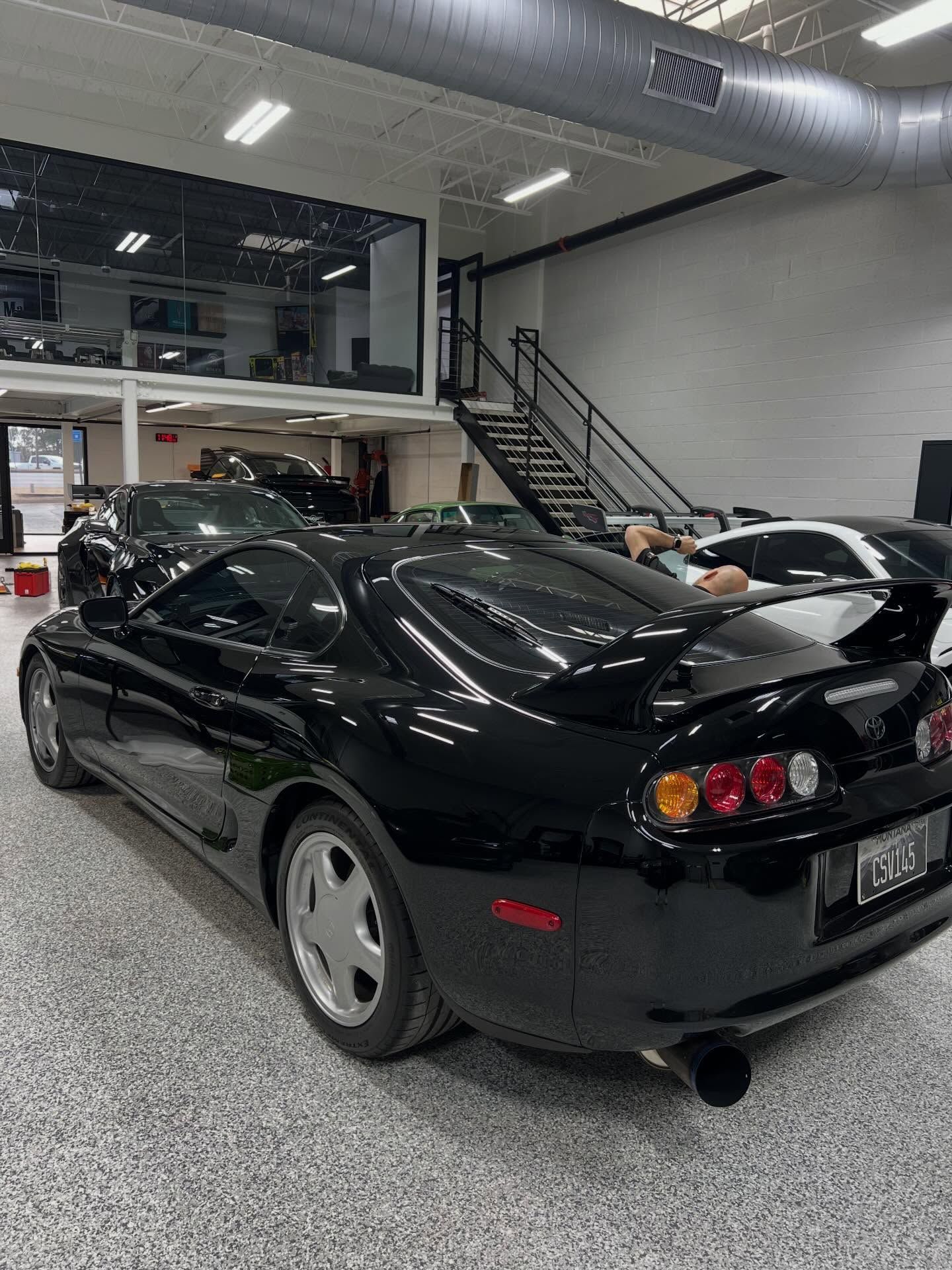 A black Toyota Supra parked in a garage with speckled flooring, near a metal staircase and other parked vehicles.