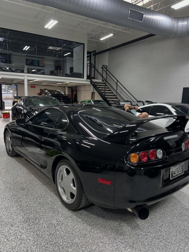 A black Toyota Supra parked in a garage with a staircase and mezzanine level in the background.