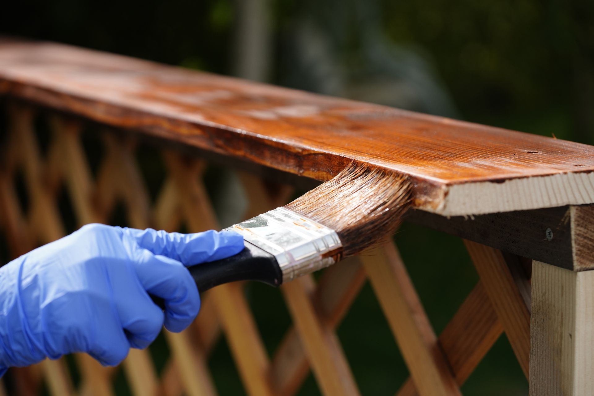 A hand in a blue glove uses a brush to apply brown wood stain to the top rail of a wooden lattice fence.