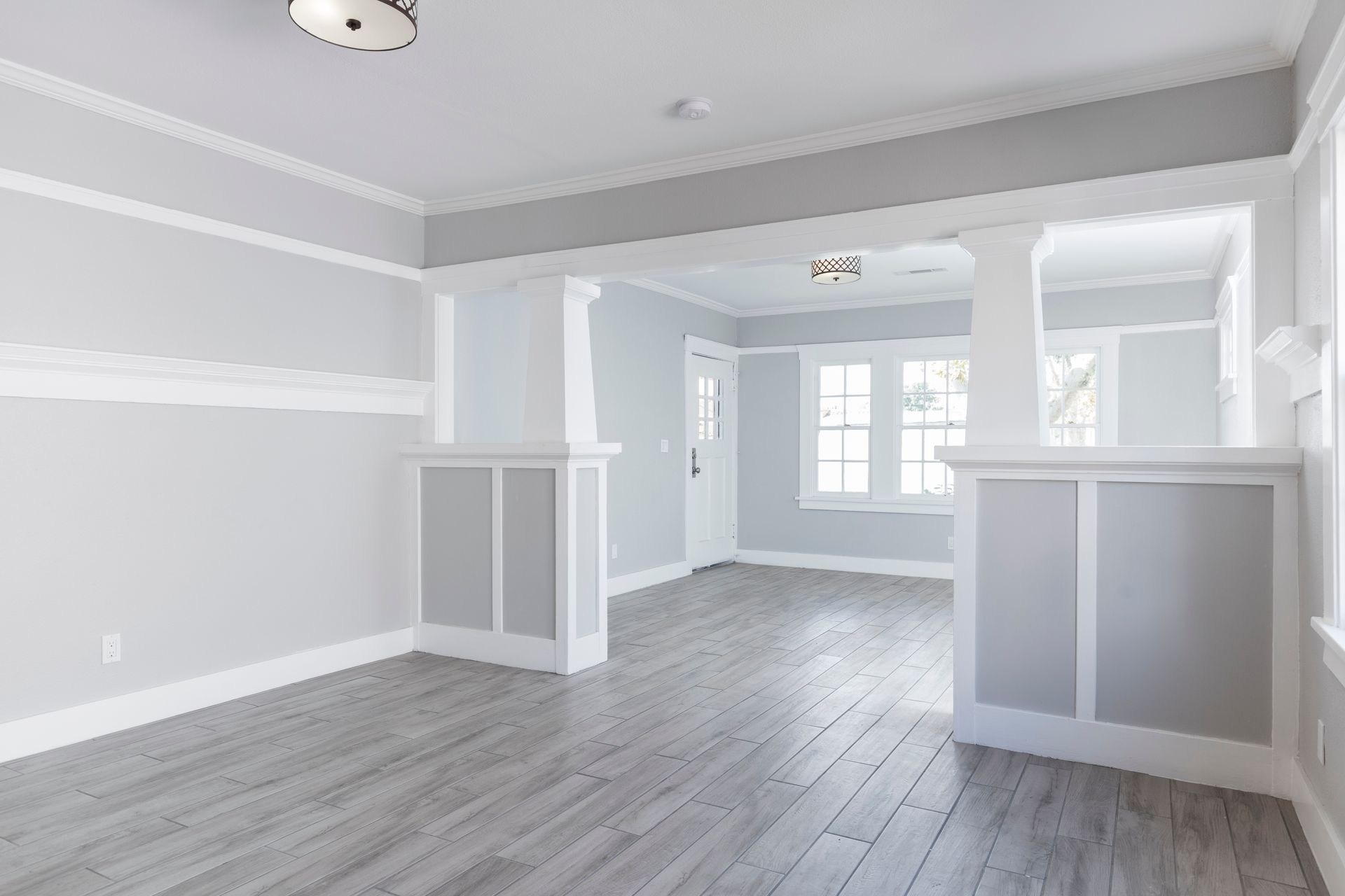 An empty, light-filled room featuring grey hardwood floors, white trim, and a columned divider between two living areas.