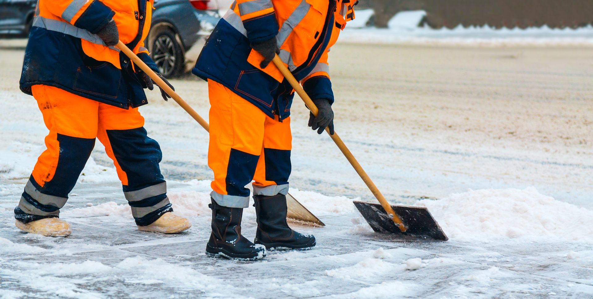 Two workers in orange and blue high-visibility gear clear snow from the ground with shovels.