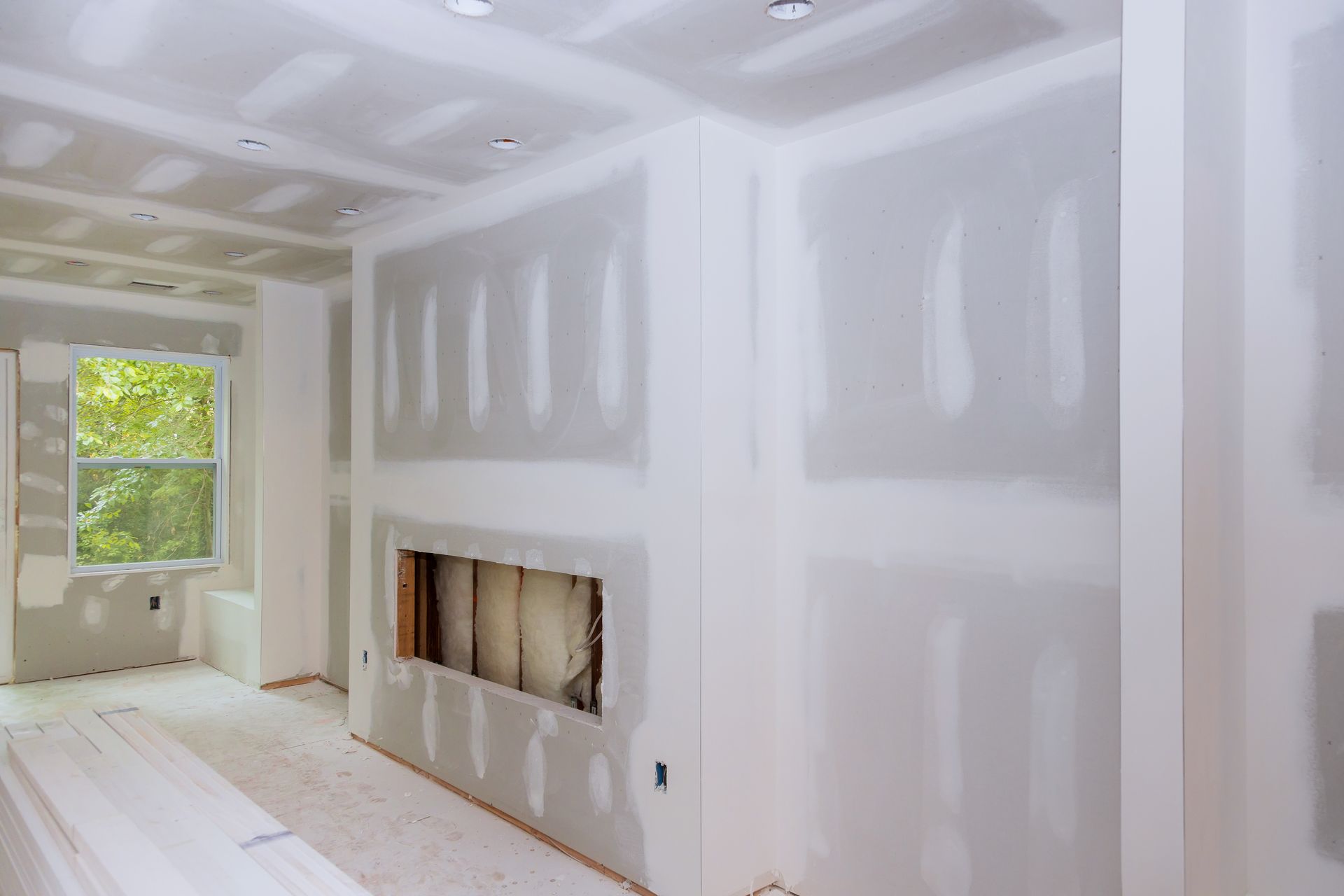 A room under construction with white drywall, taped seams, recessed lighting, and an empty fireplace insert.